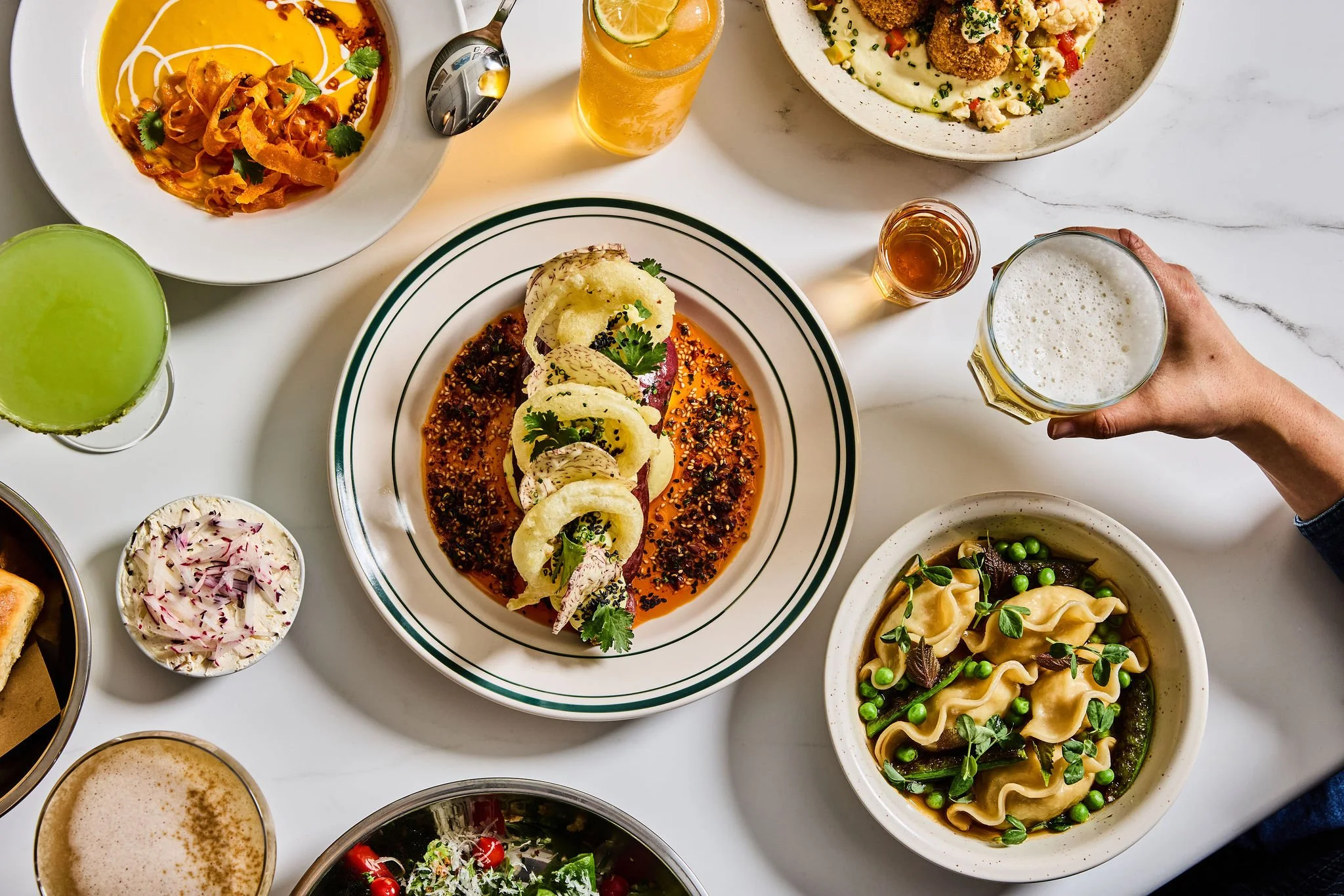 A variety of food dishes on a white table, including salads, pasta, and drinks such as beer and a citrus beverage.