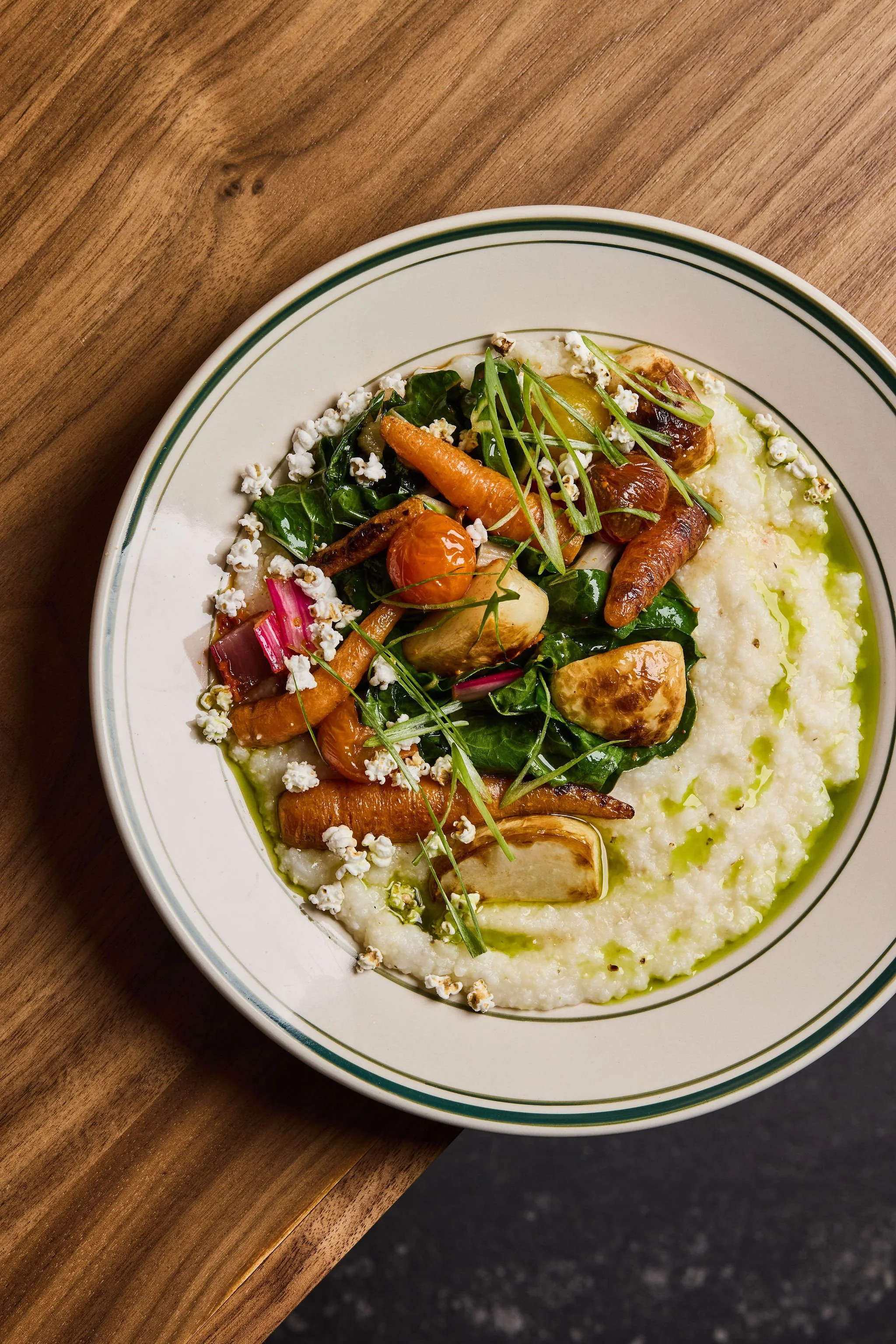 A plate of creamy mashed potatoes topped with roasted vegetables including carrots, mushrooms, cherry tomatoes, and herbs on a wooden table.