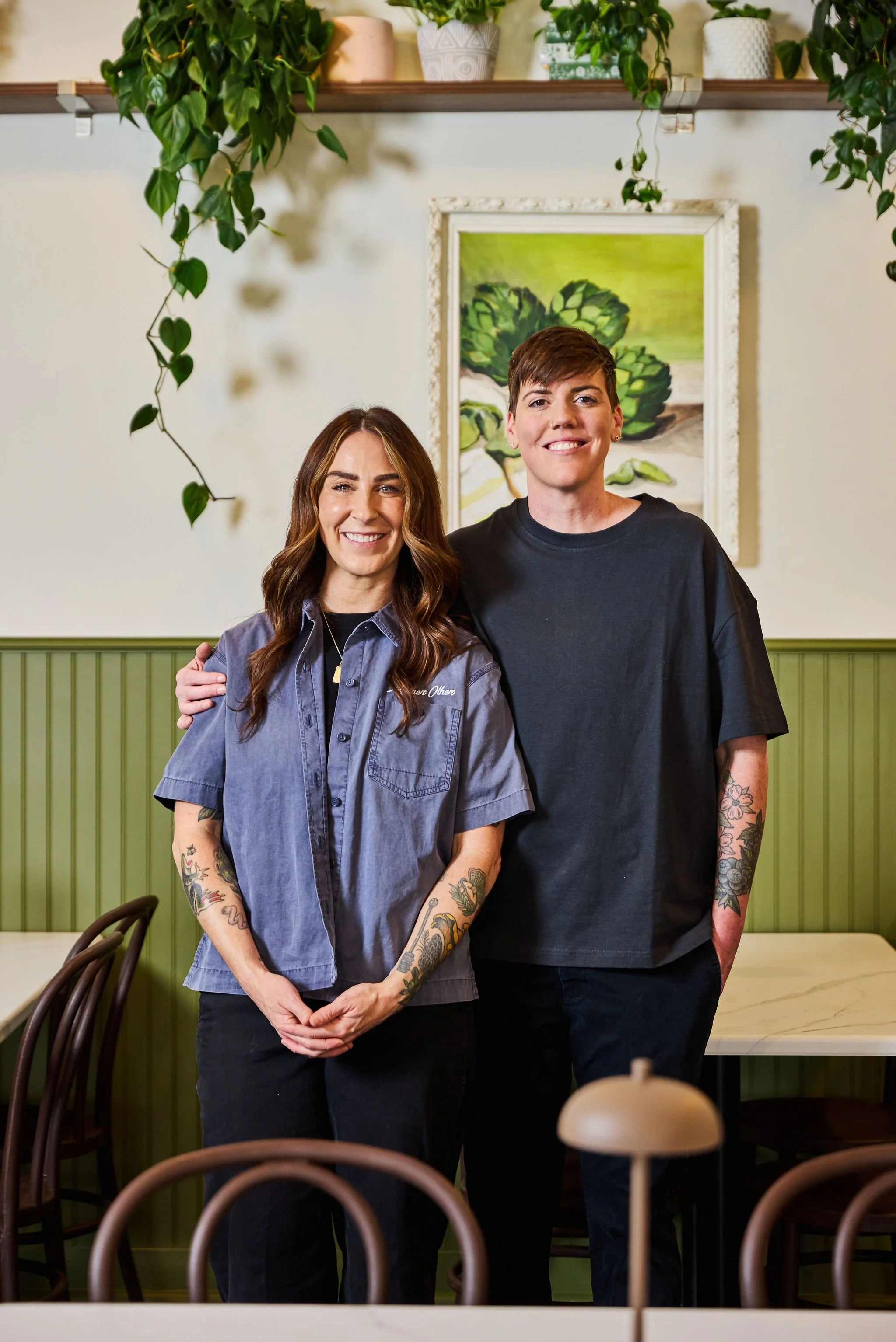 Two smiling women standing close together indoors, with their arms around each other. The woman on the left has long wavy brown hair, tattoos on her arms, and is wearing a loose denim shirt. The woman on the right has short dark brown hair, tattoos on her arms, and is wearing a black t-shirt. They are in a room with green wainscoting, a wall painting of green artichokes, and decorative plants on a shelf above them.