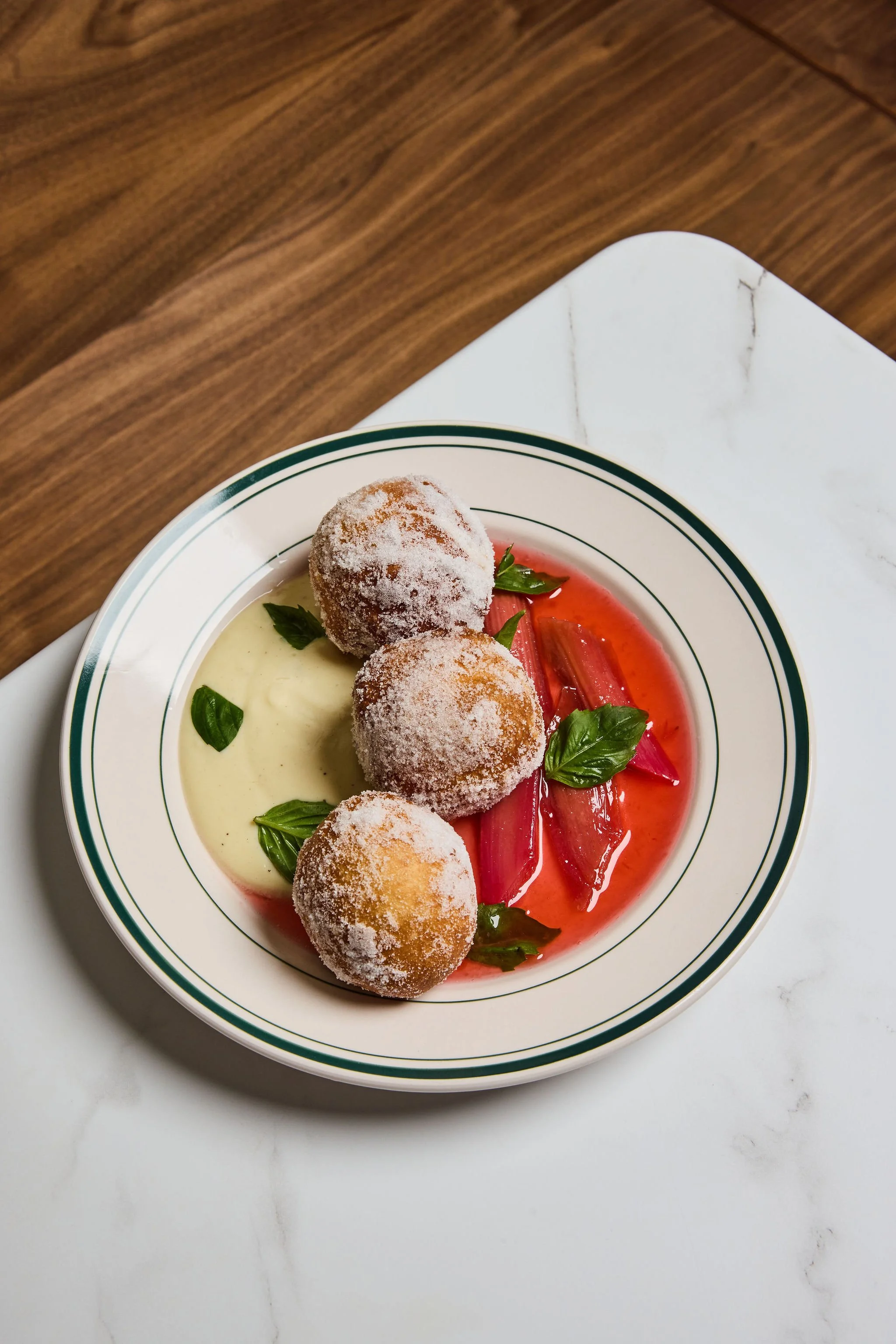 A white bowl with green stripes containing four round fried food items dusted with sugar, served with a side of pinkish-red syrup and garnished with fresh basil leaves, on a white marble surface.