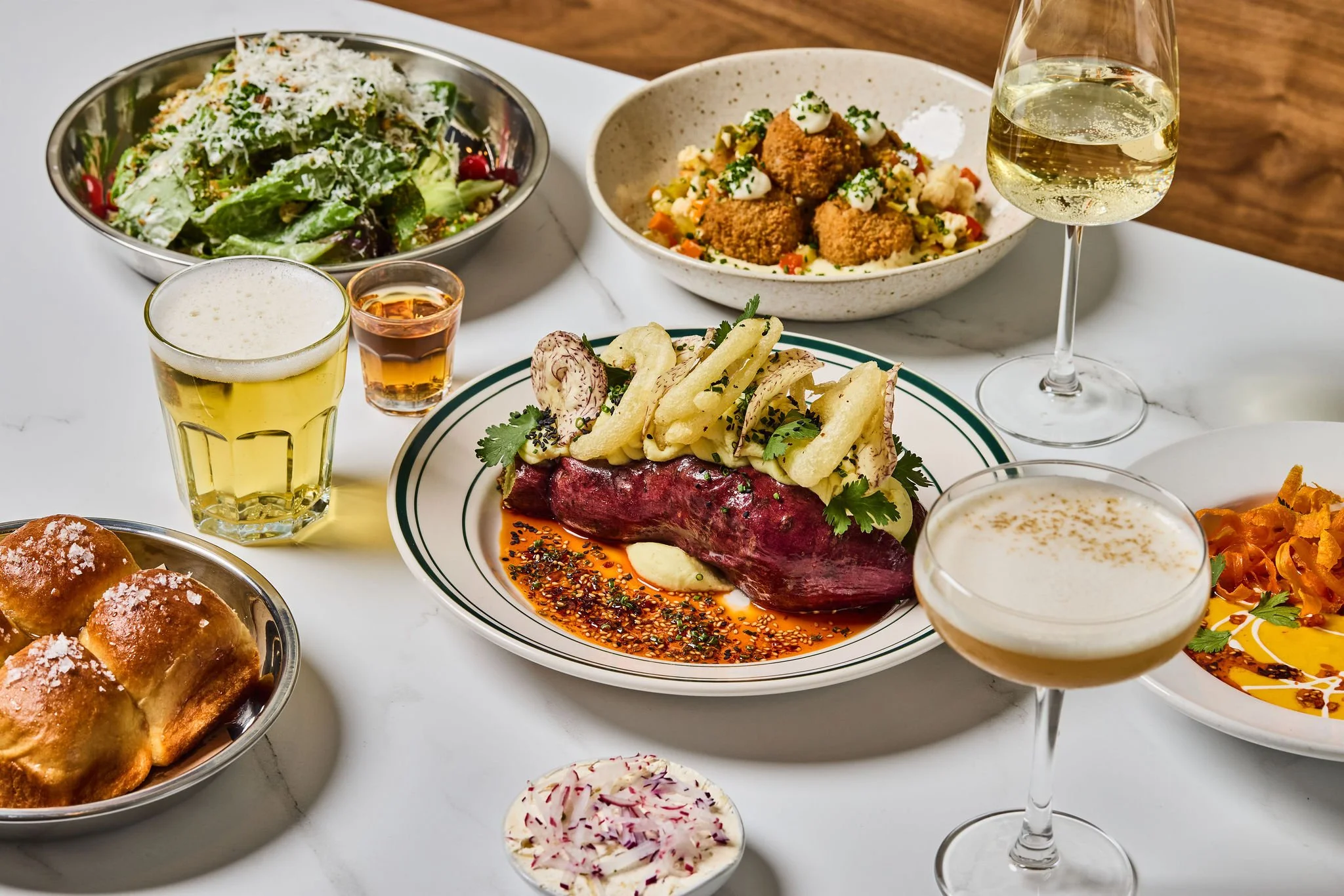 A table with various food and drinks, including a green salad, bowls of appetizer or entrée with fried items and vegetables, a glass of white wine, a glass of beer, a small shot of liquor, a plate with beef topped with pasta and herbs, and a small bowl of creamy dip or dressing.