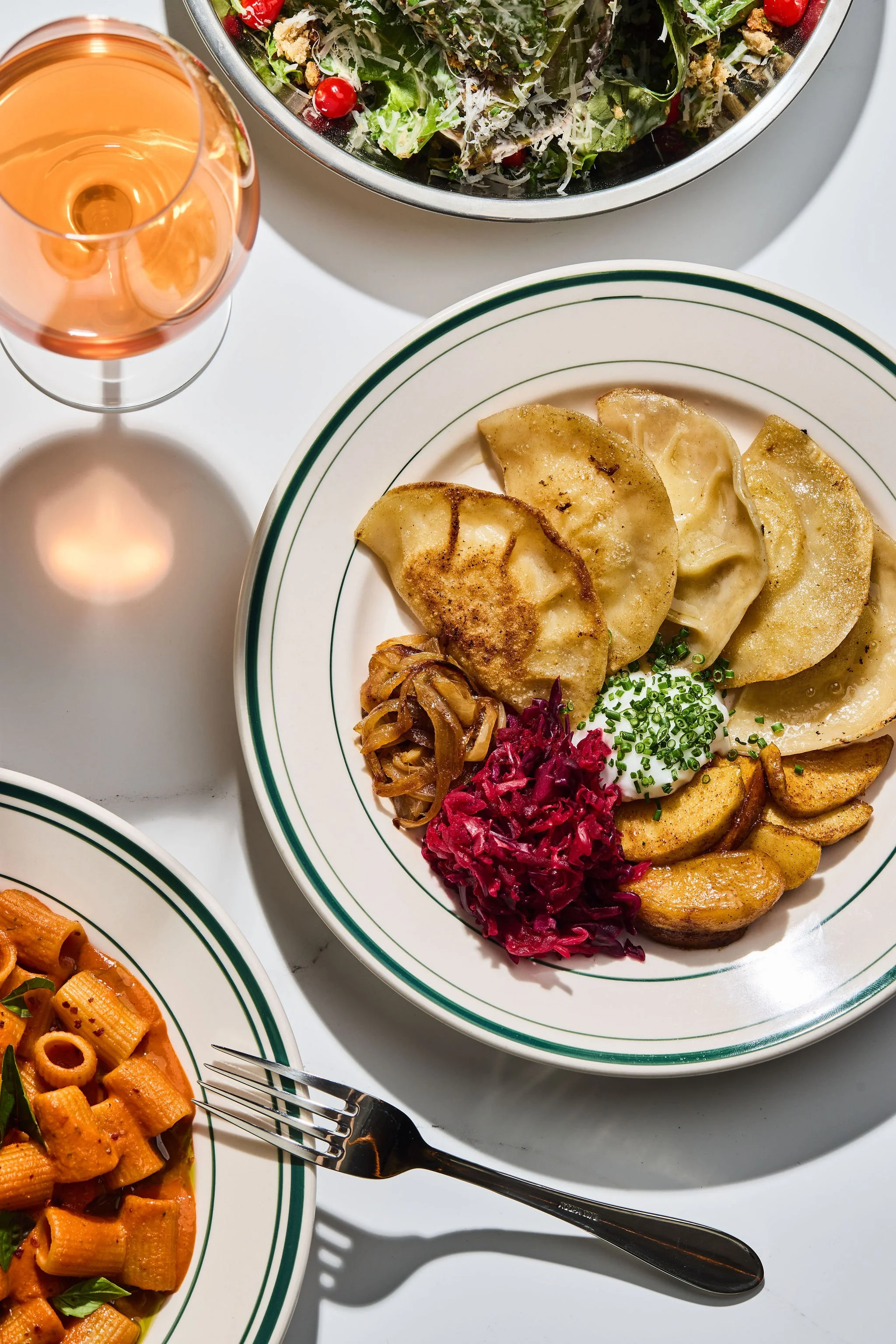 A close-up view of a dinner plate with pierogis, topped with chopped chives, served with caramelized onions and a side of shredded red cabbage. Part of another plate with pasta is visible, along with a glass of rosé wine and a bowl of salad with cherry tomatoes, greens, shredded cheese, and croutons.