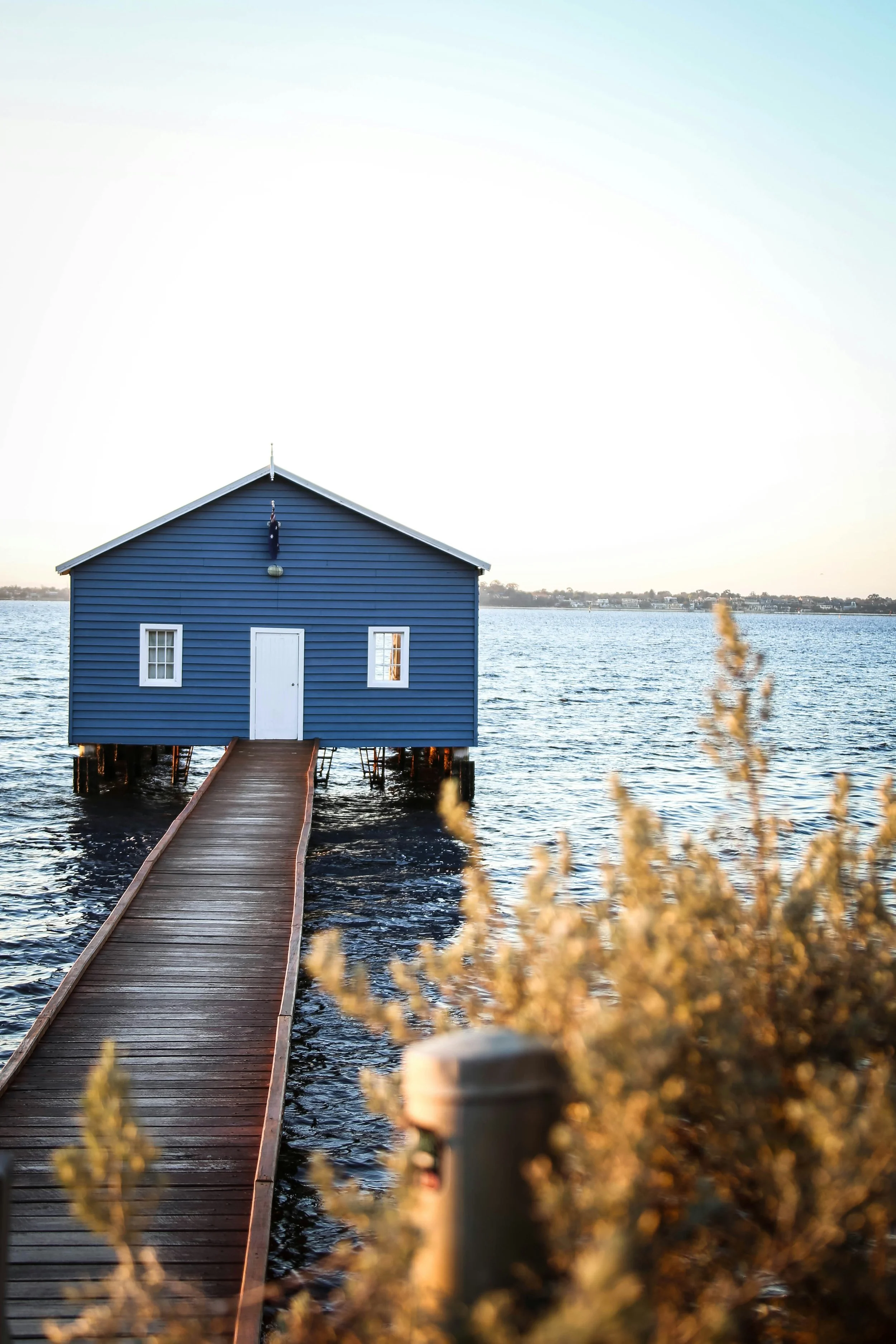 A small blue house on stilts in a body of water connected to the shore by a wooden walkway, with a distant shoreline under a light sky.