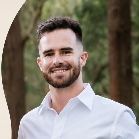 A smiling man with dark hair and a beard, wearing a white shirt, standing outdoors in a wooded area.