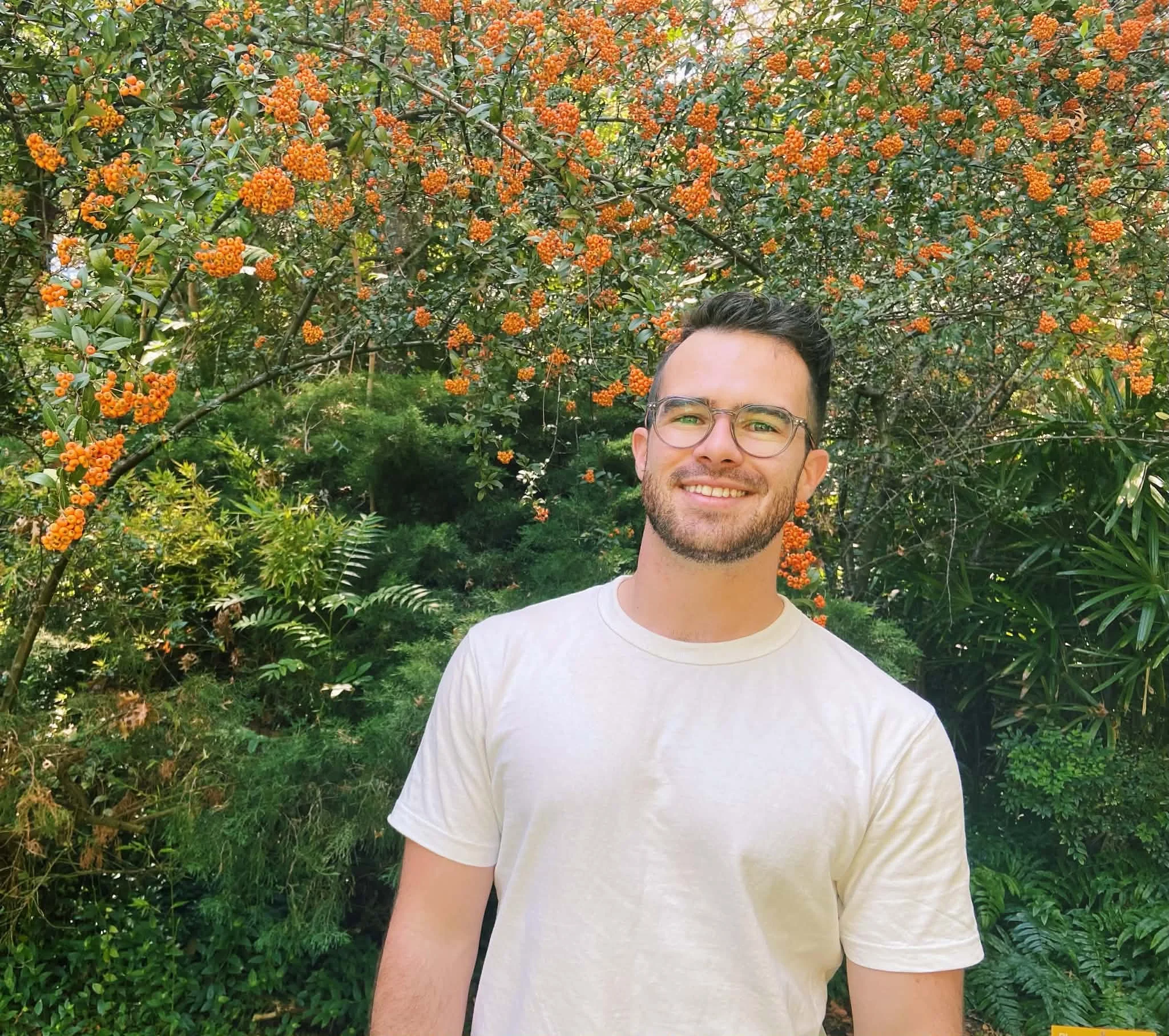 Close-up of a smiling man with glasses and a beard, wearing a white t-shirt, standing outdoors in front of green foliage and orange berries on a tree.