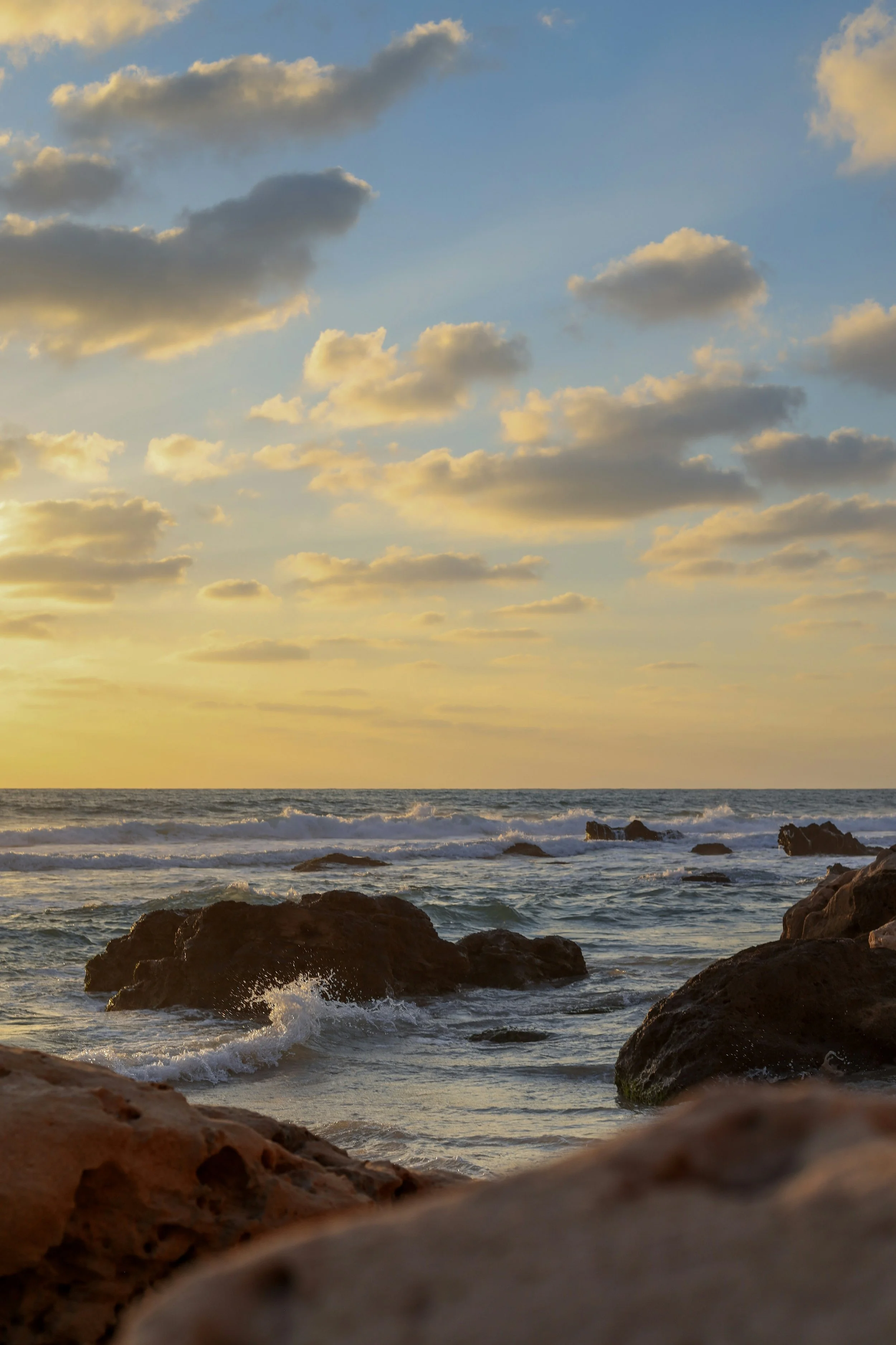 Sunset over the ocean with clouds and rocks in the foreground.