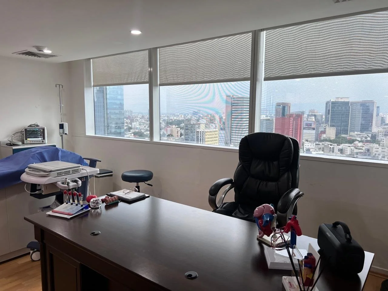 Office with a large wooden desk, black leather chair, hospital equipment, and a city view through tall windows.