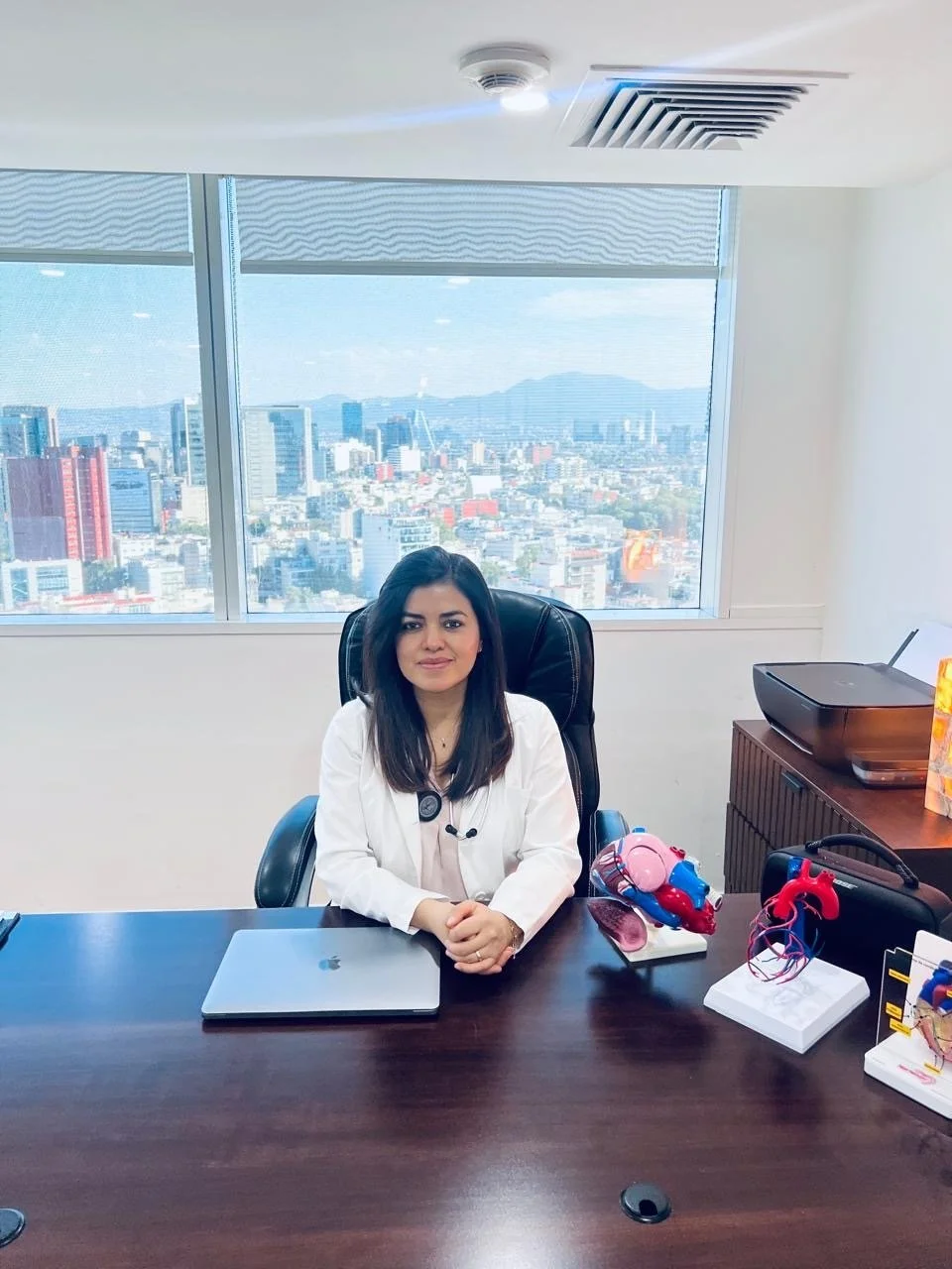 A woman with dark hair sitting at a large wooden desk in an office with a city skyline visible through the window behind her, wearing a white coat.