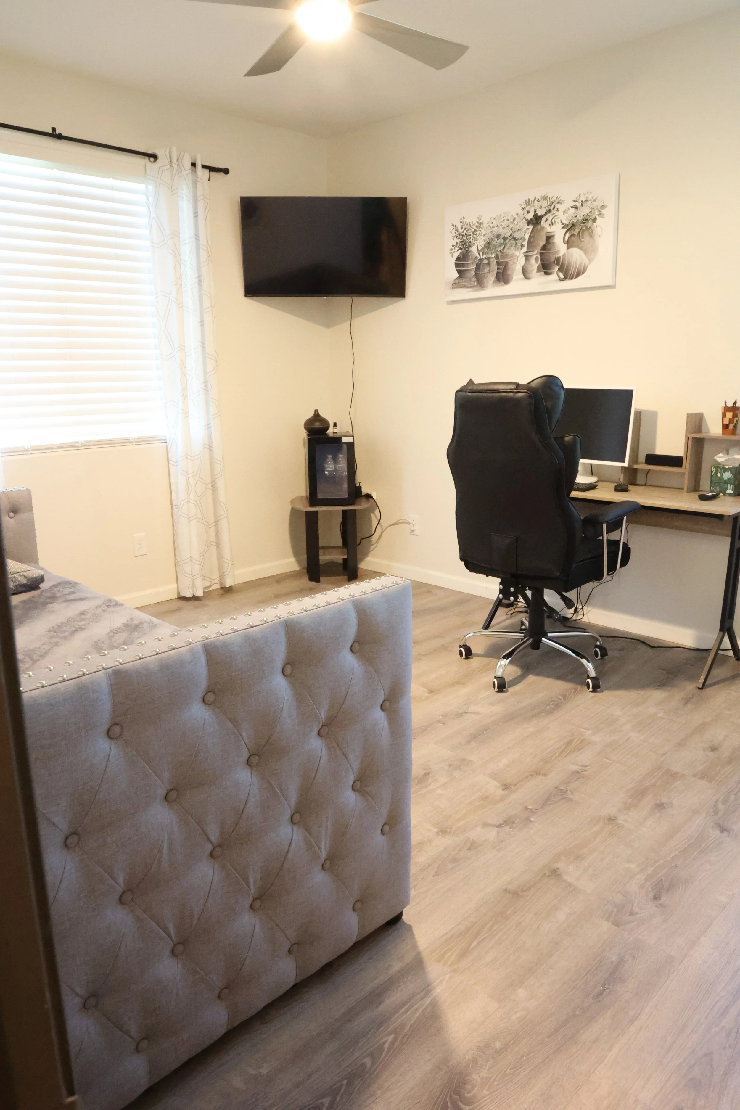 Living room with a beige tufted couch, a wall-mounted television, a black office chair at a desk with a computer, a window with blinds and curtains, a wall art of vases, and a small side table with digital devices.