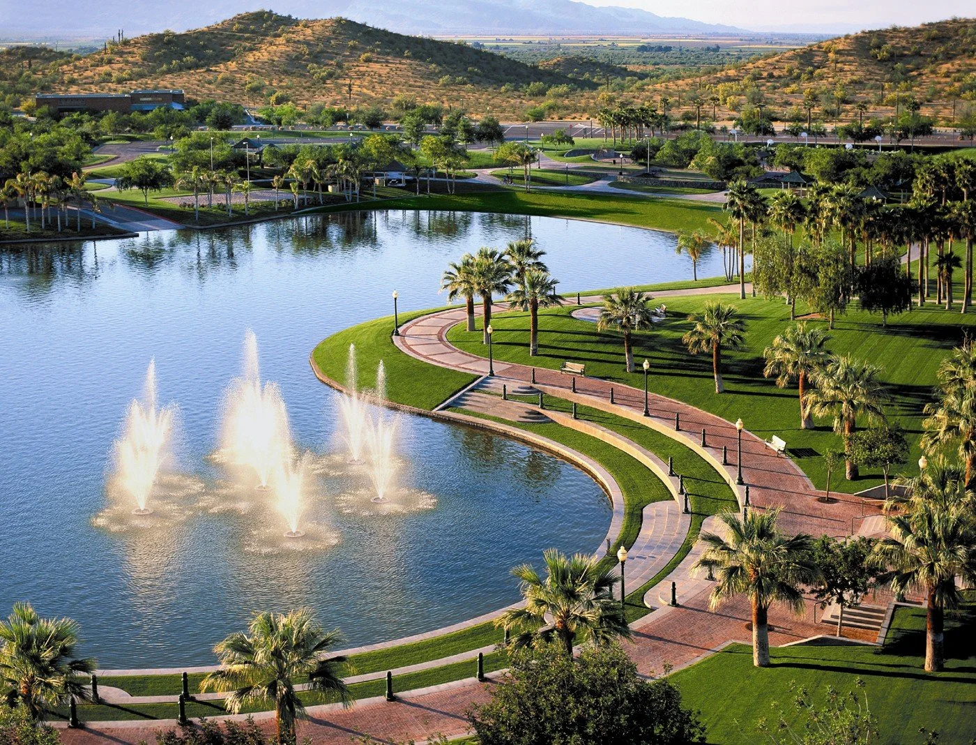 A scenic park with a large pond featuring four fountains, walking paths, numerous palm trees, benches, and street lamps, with hills and a desert landscape in the background.