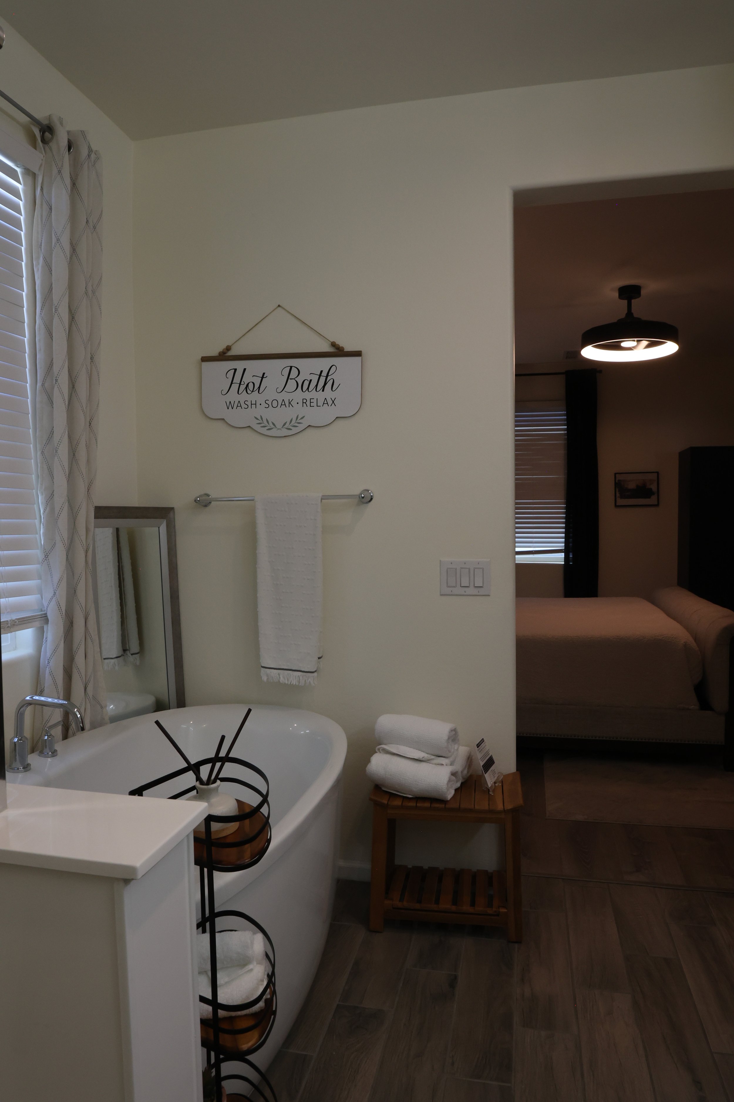 A cozy bathroom with a vintage-style bathtub and a sign that reads 'Hot Bath Wash Soak Relax'. The space includes neatly stacked towels on a small wooden stand, a towel rack, and a window with curtains. Adjacent to the bathroom, a bedroom with a bed 