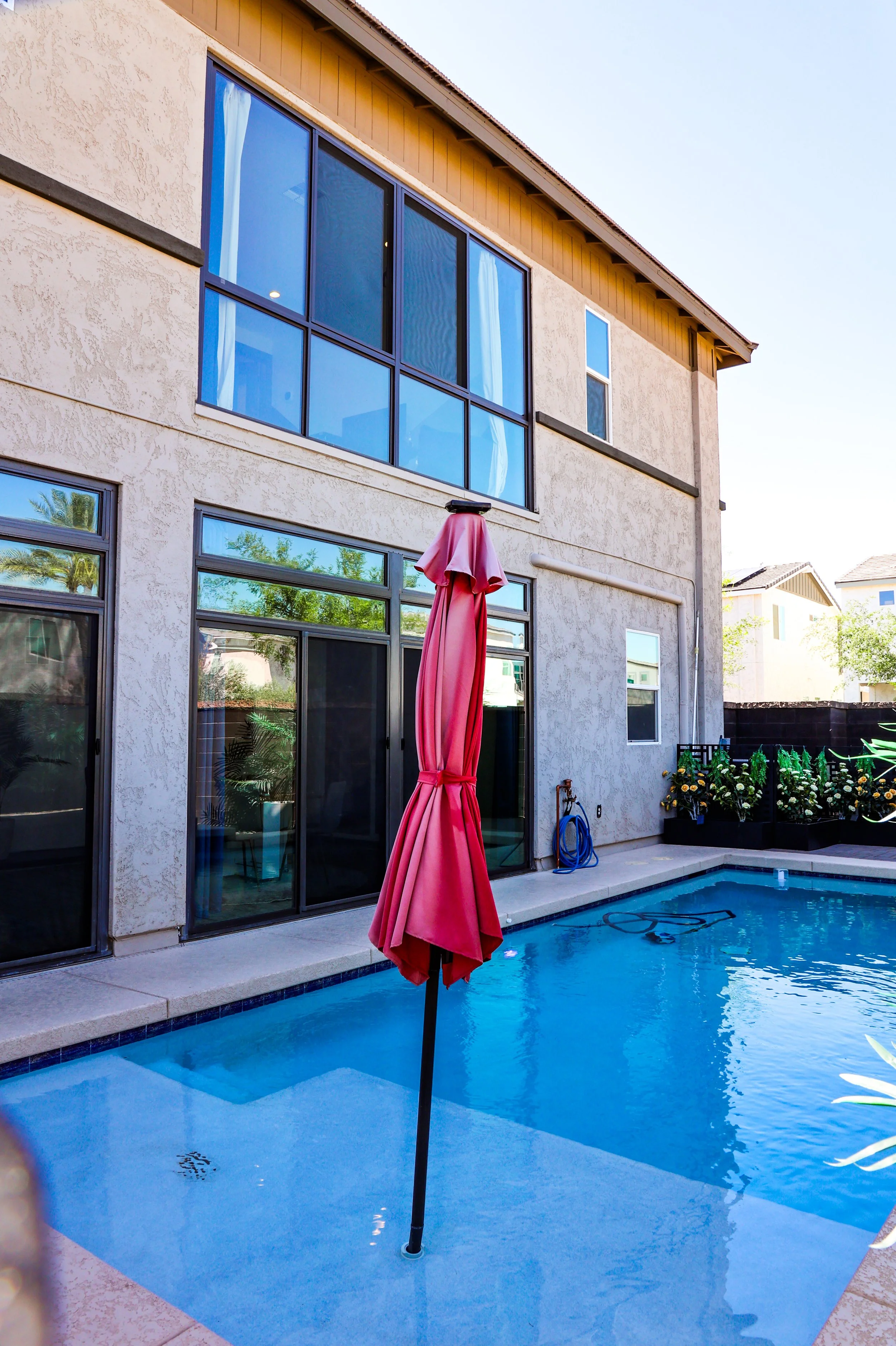 A house with large glass sliding doors and windows, a pink pool umbrella, a swimming pool, and plants in the backyard.