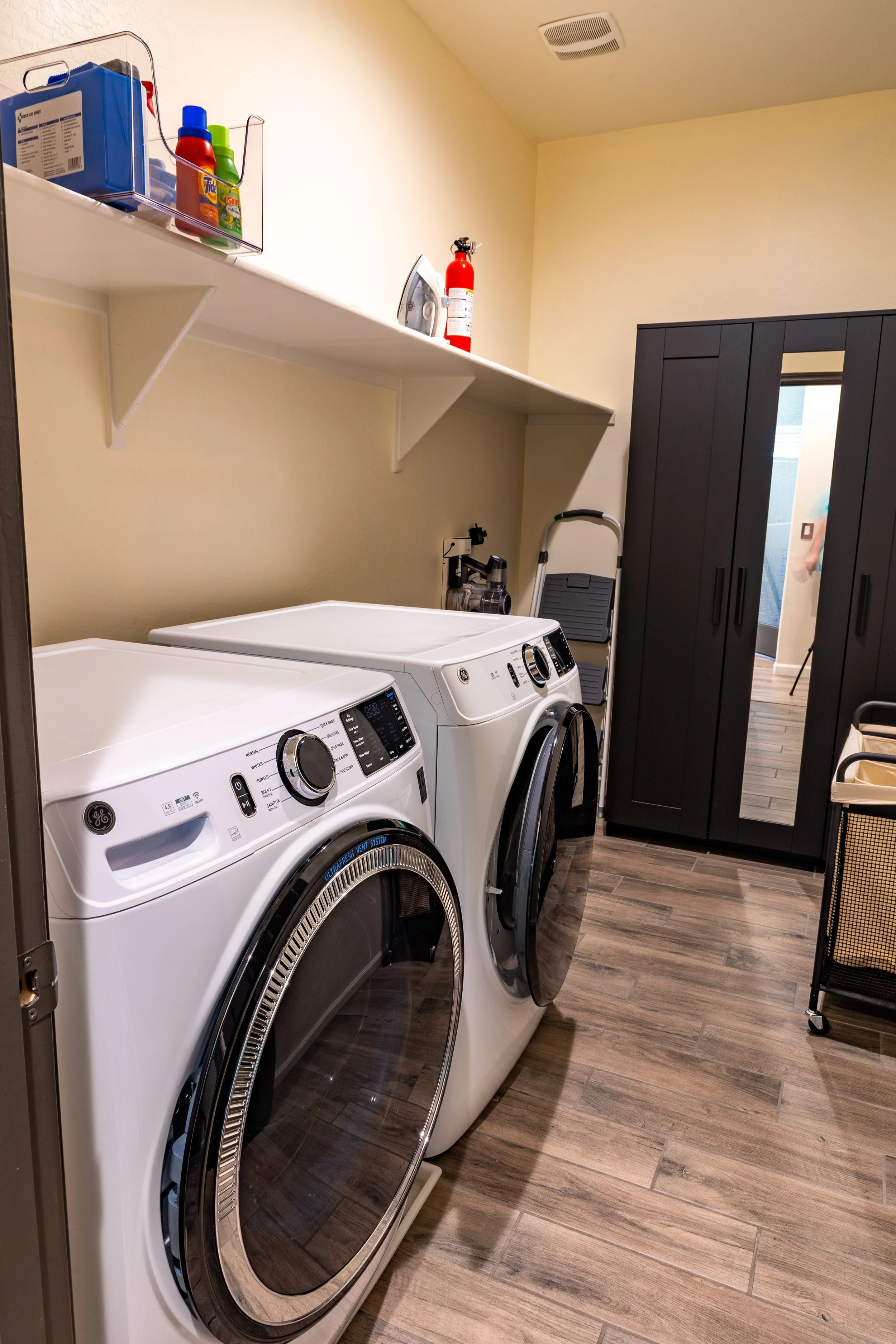 A laundry room with a pair of white front-loading washing machines and a dark brown cabinet with a mirror door. There is a shelf above the machines with laundry supplies and a fire extinguisher. A small vacuum cleaner and a stepladder are in the corn
