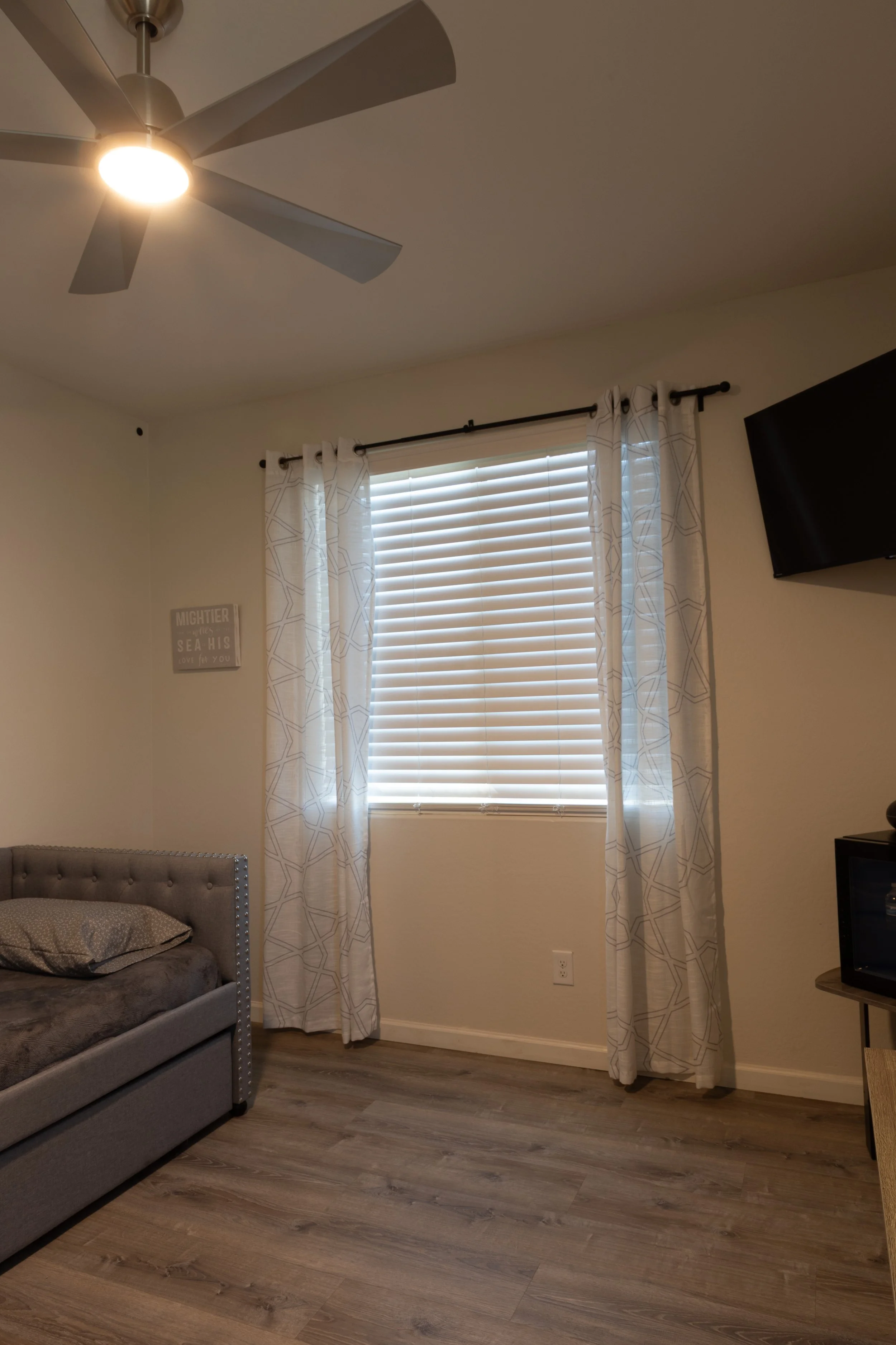 A corner of a bedroom showing a window with blinds and white patterned curtains, a gray upholstered bed with a pillow, a wall decoration, a wall-mounted TV, and a ceiling fan with lights.