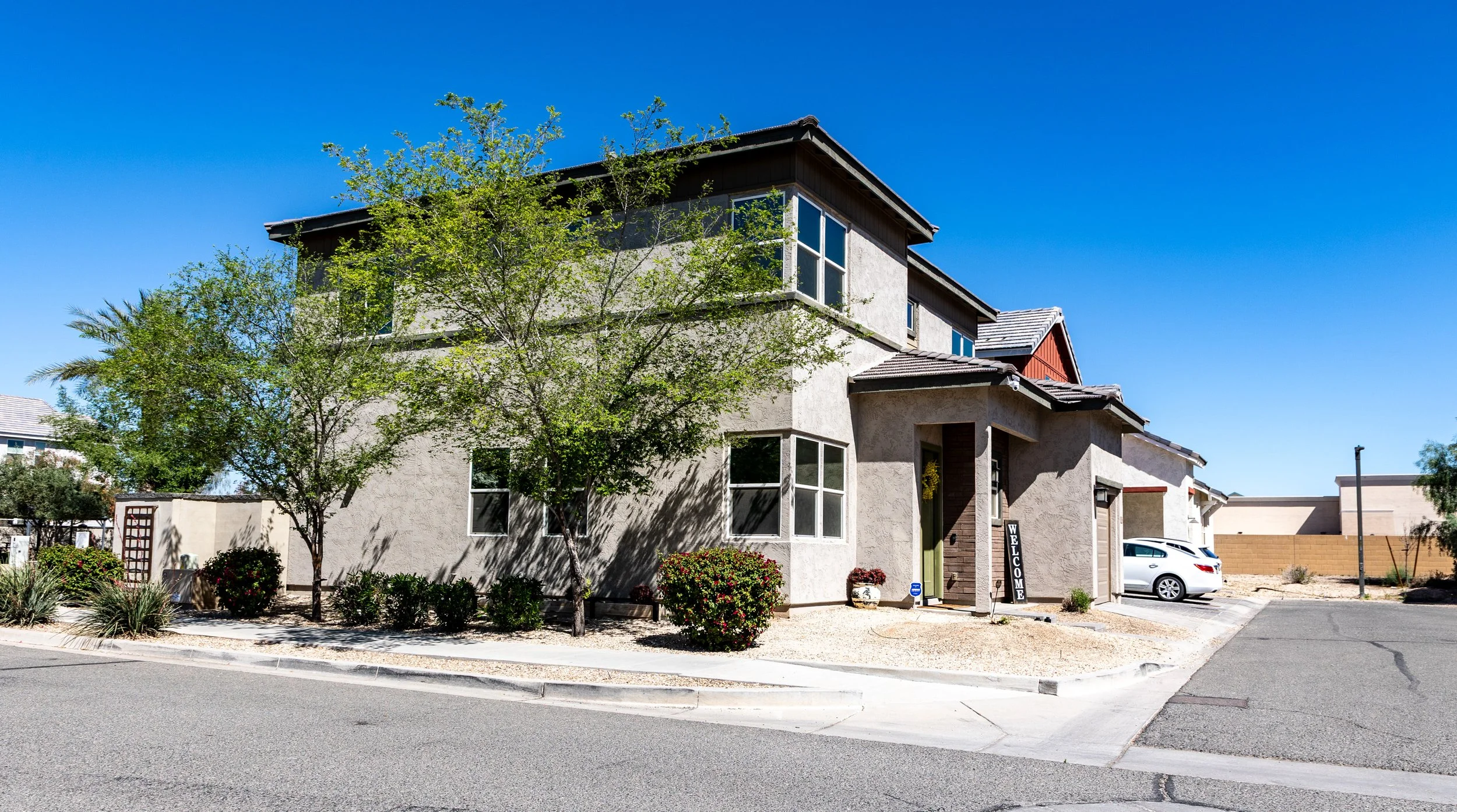 A modern multi-story house with white stucco walls, multiple large windows, and a combination of flat and sloped roofs. There are trees and bushes in the front yard, a sidewalk, a small parking lot with a white car, and a blue sky in the background.