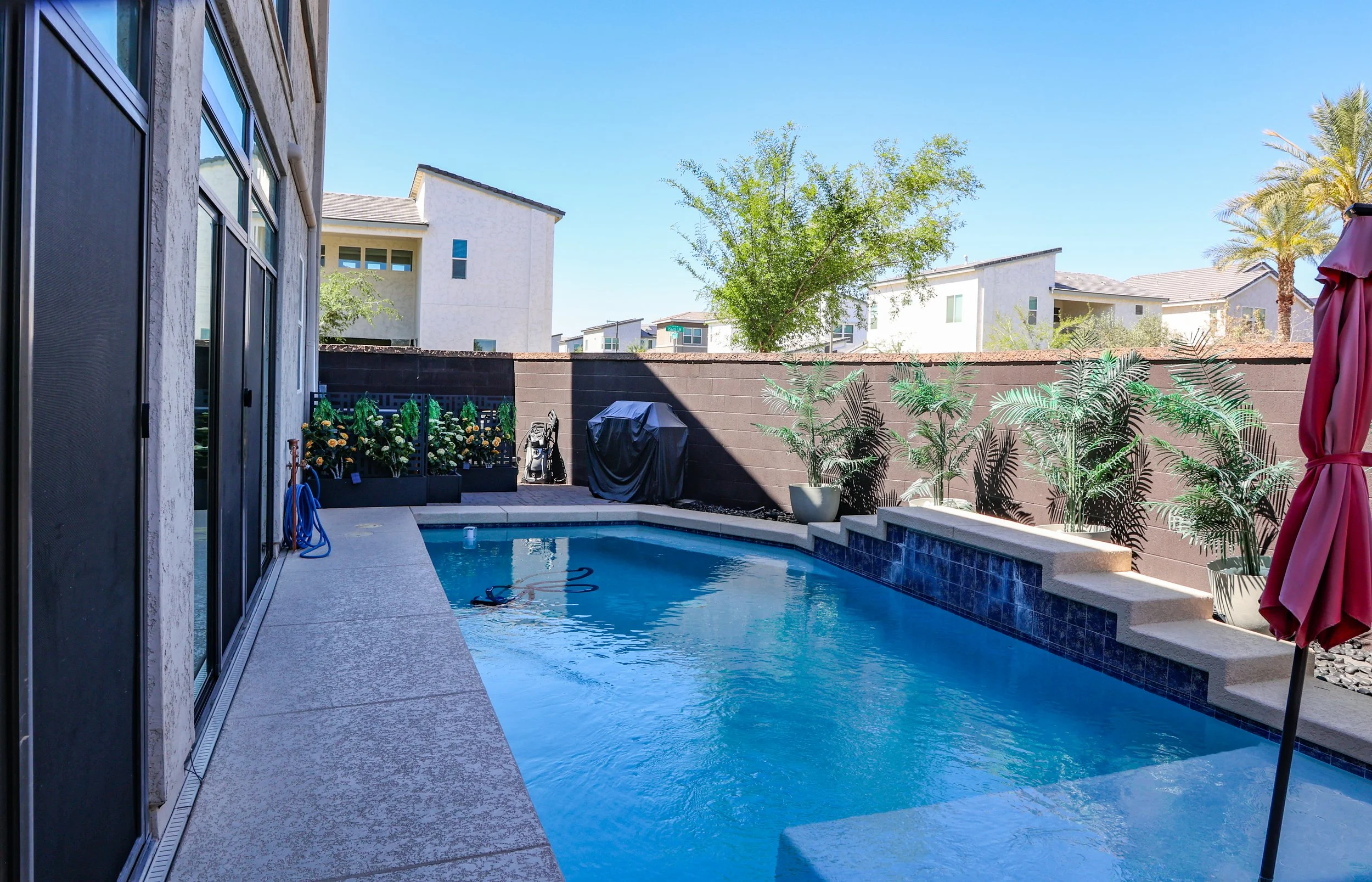 Backyard with a small swimming pool, umbrellas, potted plants, and a brown privacy wall, with house and trees visible in the background.