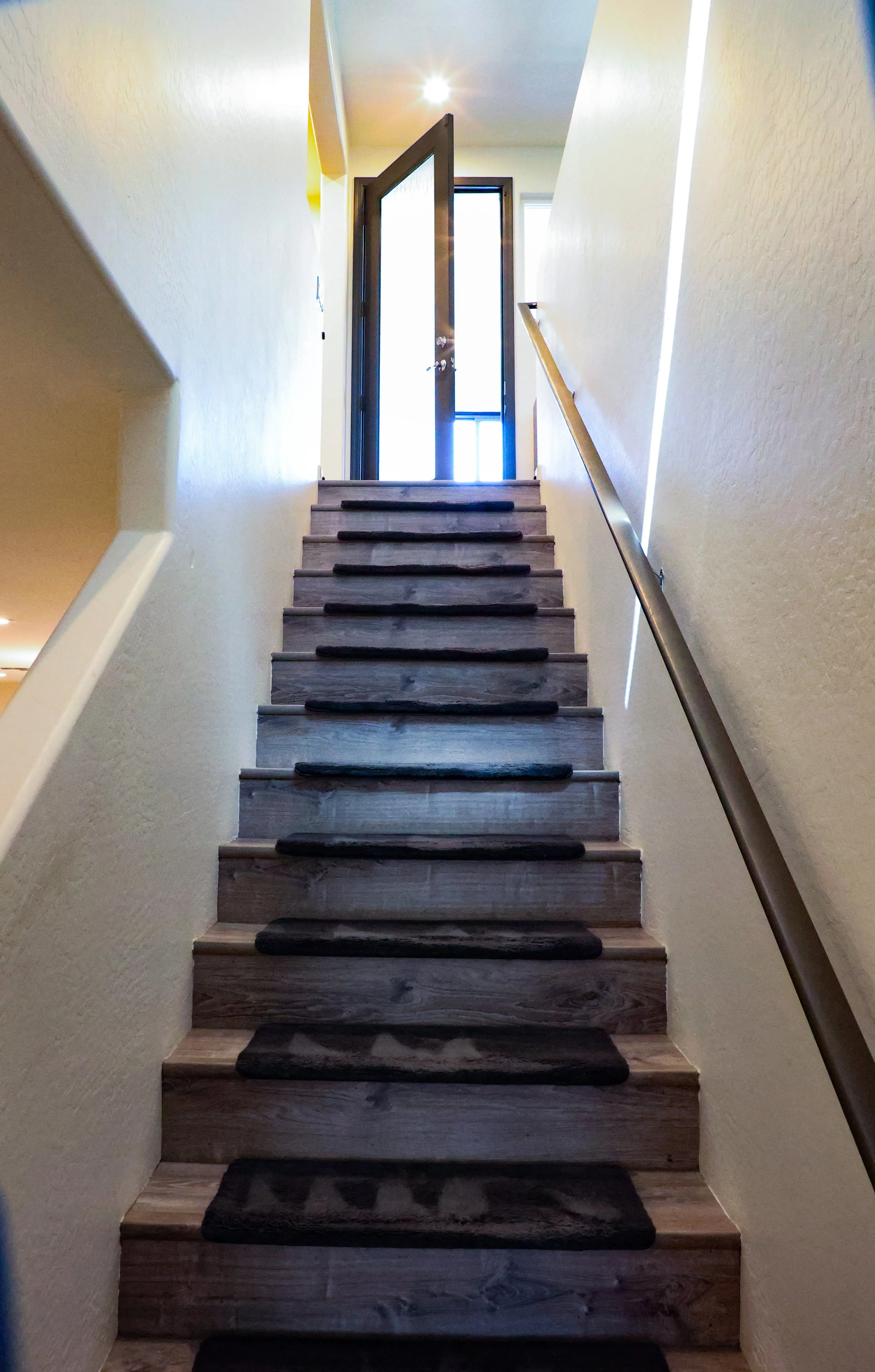 Wooden staircase with dark tread mats, beige walls, a handrail on the right side, leading up to an open front door with light flooding in.