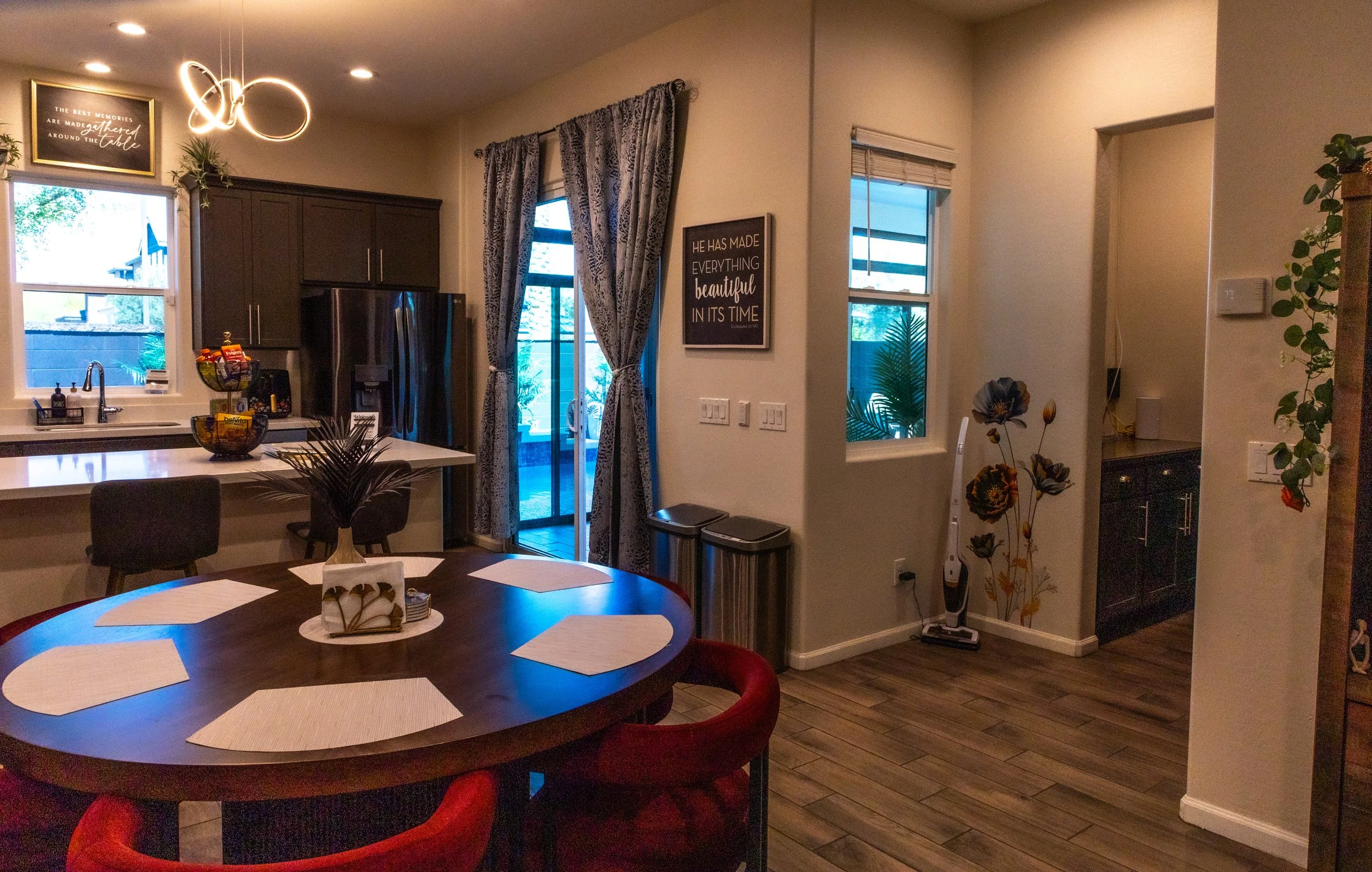 A cozy kitchen and dining area with a round wooden table surrounded by red chairs, decorated with placemats and a napkin holder. The kitchen has dark cabinets, stainless steel appliances, and a window above the sink. There are curtains on the sliding