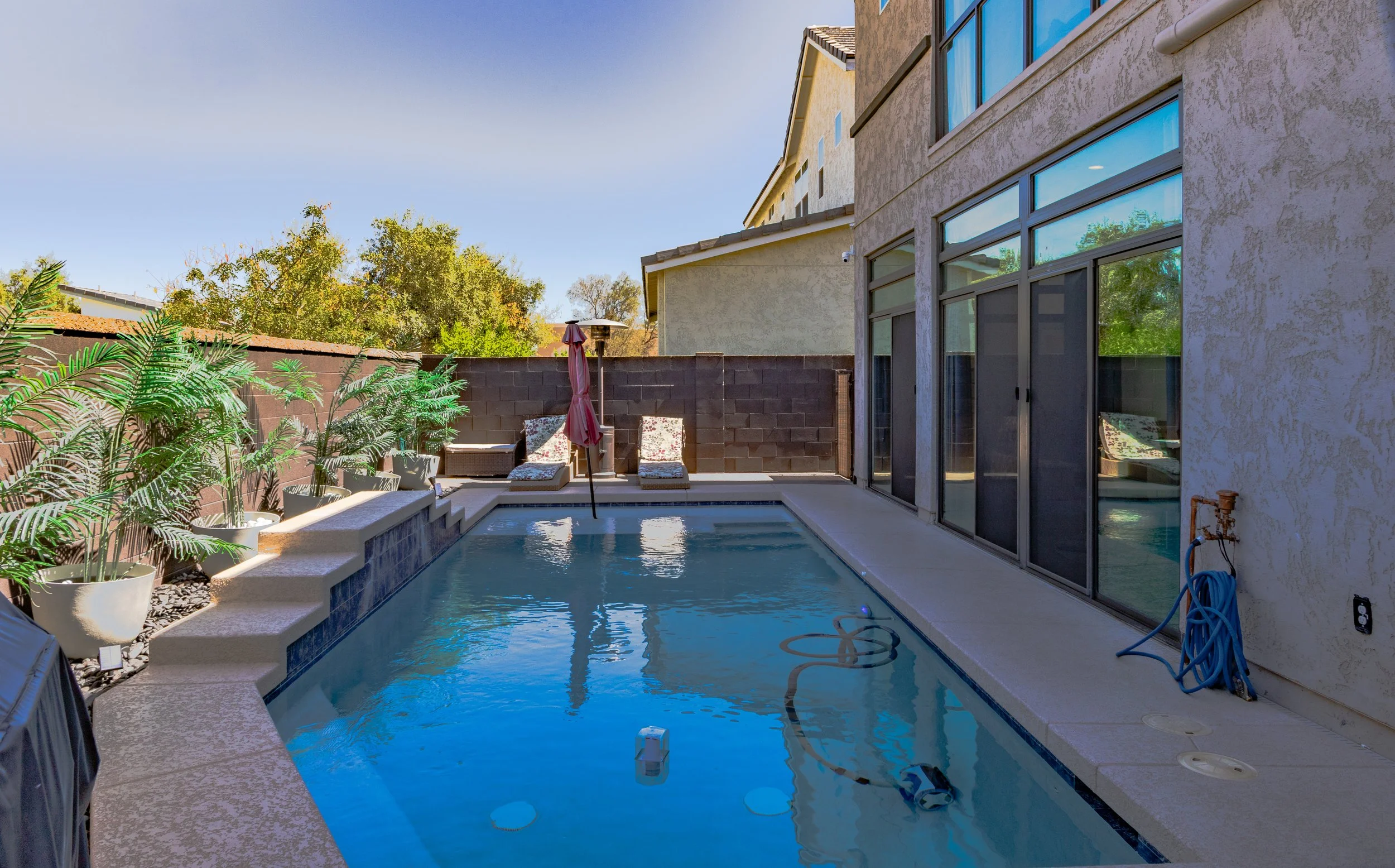 A backyard swimming pool area with lounge chairs, an umbrella, and potted plants, surrounded by a privacy wall next to a beige stucco house with large glass sliding doors.