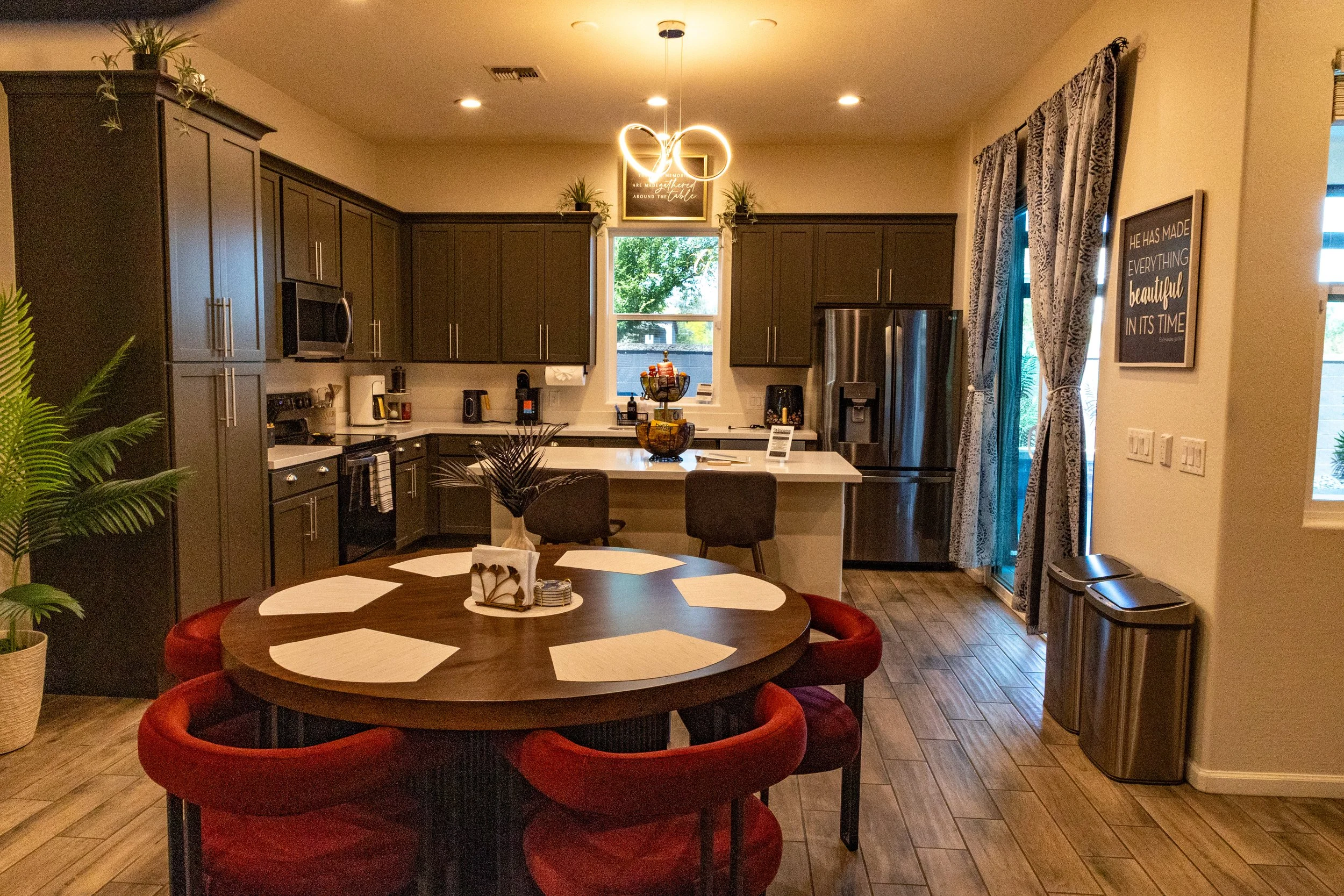 Modern kitchen and dining area with dark cabinets, stainless steel appliances, a round wooden table with red chairs, decorative plants, and window with curtains.