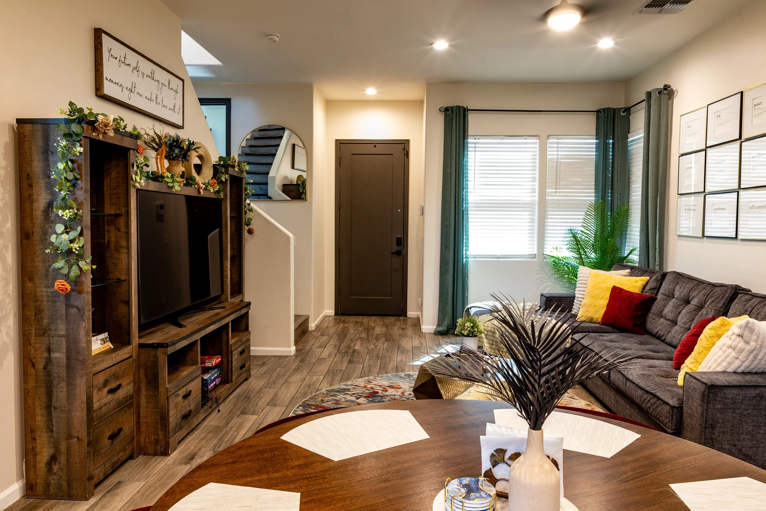 Living room with a brown sofa featuring yellow, red, and beige pillows, a wooden entertainment center with decorations, a mirror on the wall, large windows with blinds and green curtains, and various plants. A round wooden dining table with a vase an