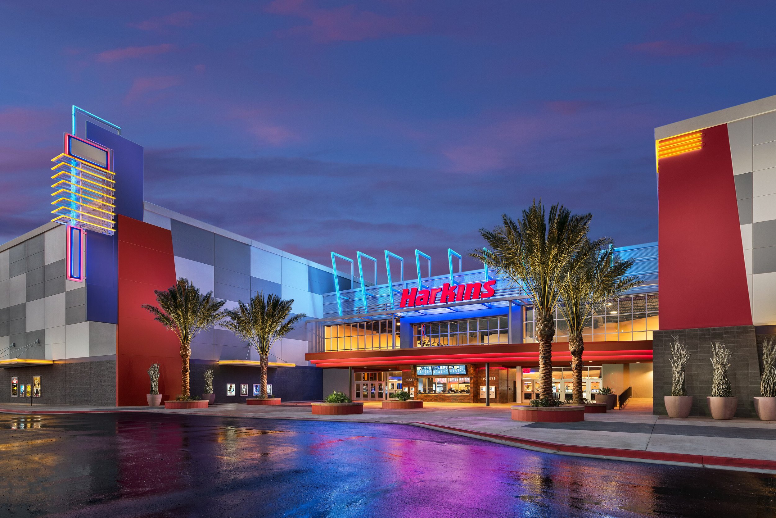 Nighttime exterior view of a modern retail store with neon lights and palm trees in front. The store has the name 'Harkins' displayed above the entrance, with colorful lighting accents on the building.