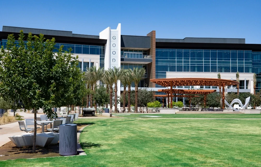A modern building with large glass windows and a sign that reads 'GODDARD' on a white vertical panel. The foreground features a park with green grass, trees, a wooden pavilion, and seating areas, under a clear blue sky.