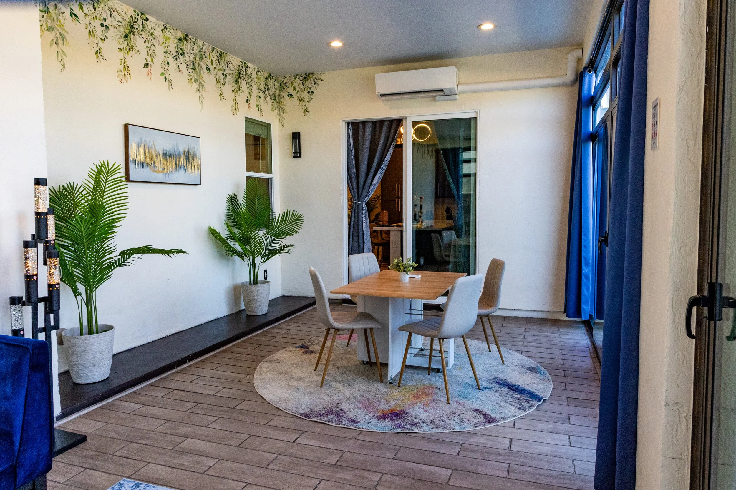 A cozy indoor dining area with a wooden table, four white chairs, a round colorful rug, potted plants, a wall painting, and curtains by a sliding glass door leading outside.