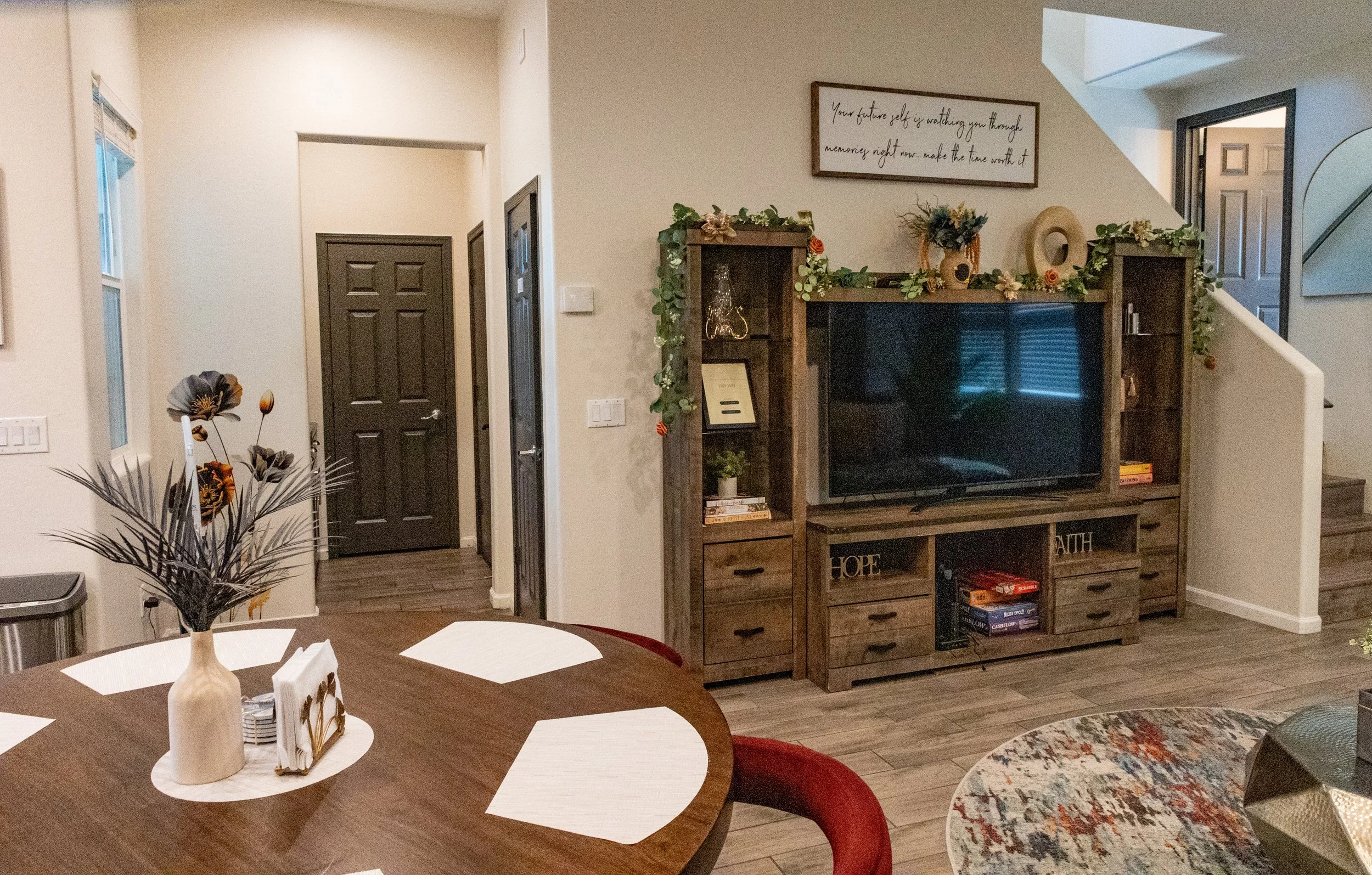 A living room with a wooden entertainment center holding a television, decorated with greenery and flowers. A framed quote hangs above, and a staircase is visible on the right. In the foreground, a dining table is set with napkins, a vase with flower