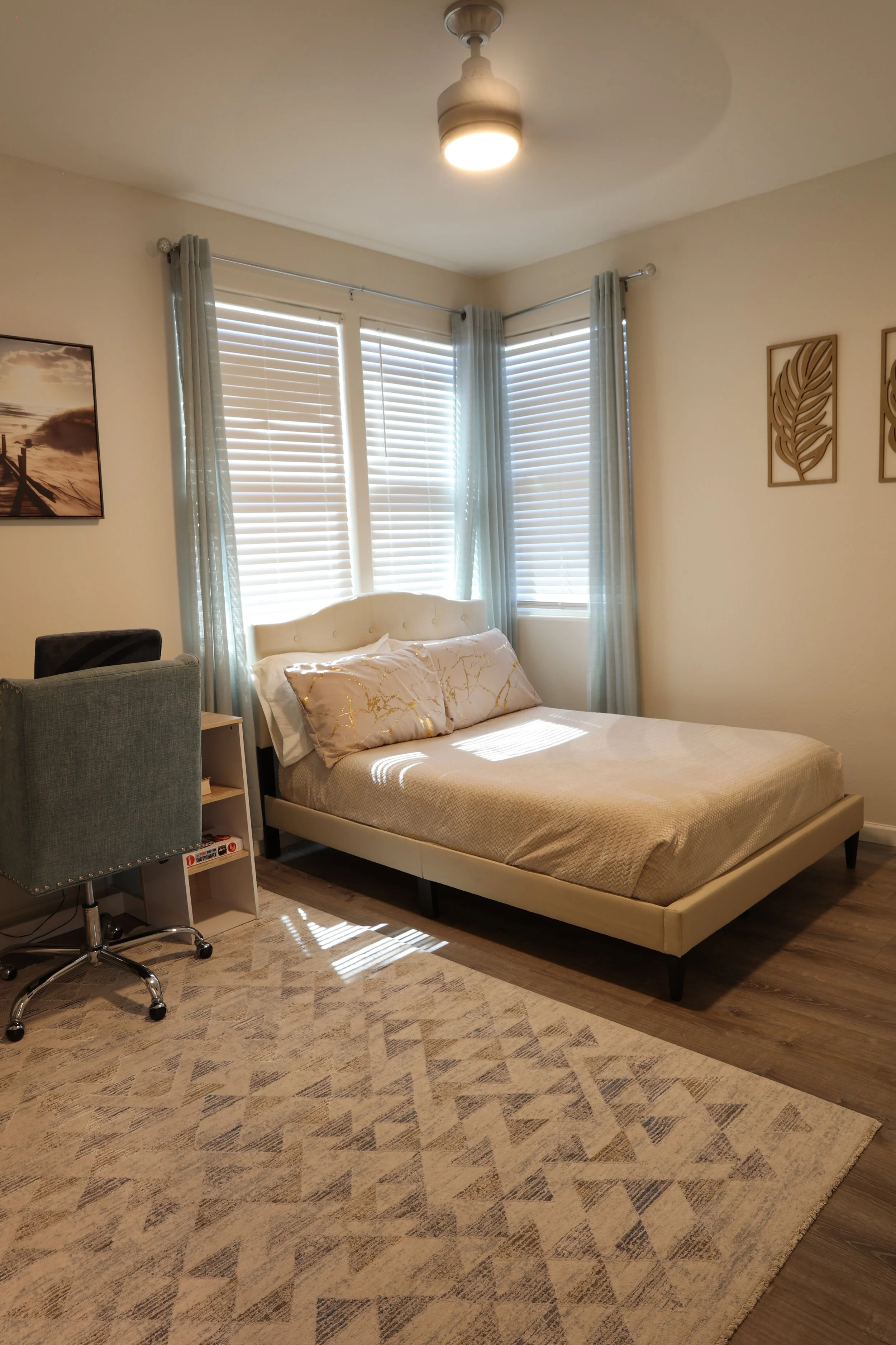 A cozy bedroom with a bed, a small desk with a chair, and large windows with blinds and light blue curtains. The room has wall art and a patterned rug on the wooden floor.