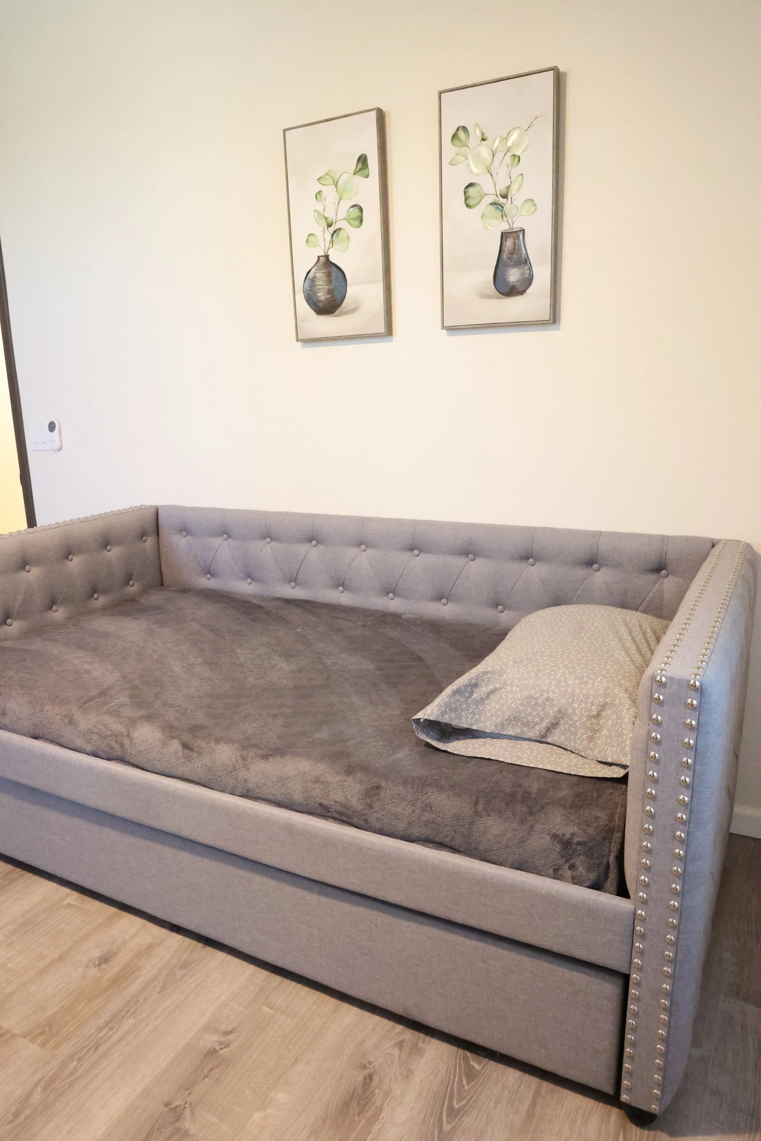 A gray upholstered sofa or daybed with a curved back and button tufting along the top edge. There is a decorative pillow with a light pattern resting on the right arm of the furniture. Two framed floral paintings featuring white and green leaves in d