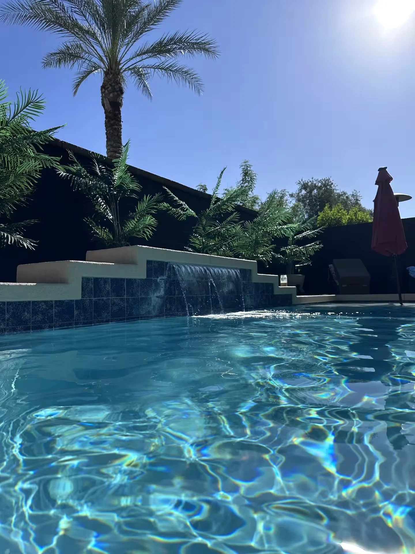 A backyard swimming pool with clear blue water, surrounded by lush green plants and trees, including a palm tree. There is a waterfall feature at the edge of the pool, and a red patio umbrella is visible on the right side under the bright, sunny sky.