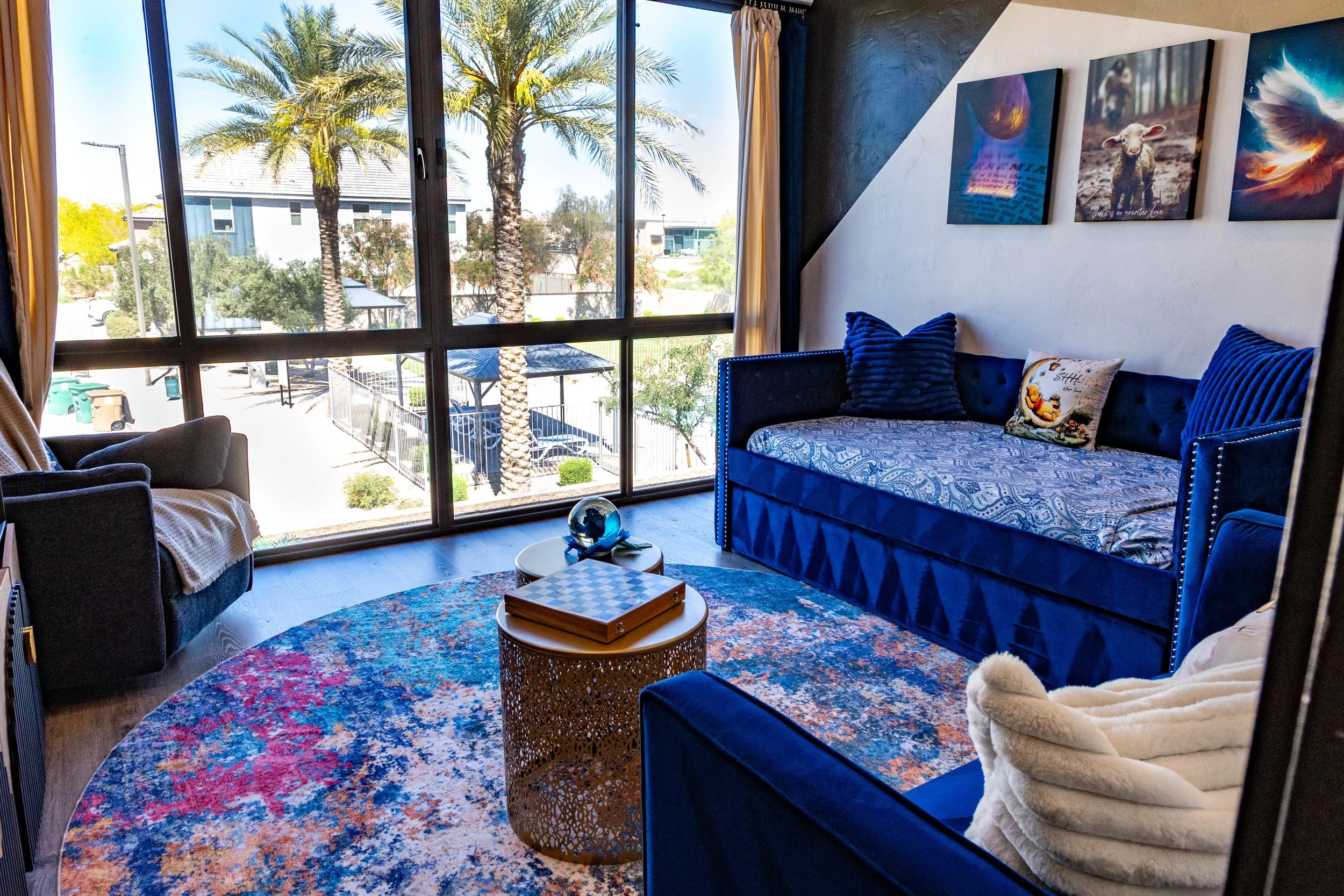 Living room with large windows showing outdoor palm trees and a street with buildings, including parked cars. Inside, there is a blue tufted sofa with pillows, a round coffee table with a chessboard and wooden box, colorful rug, armchair with throw b