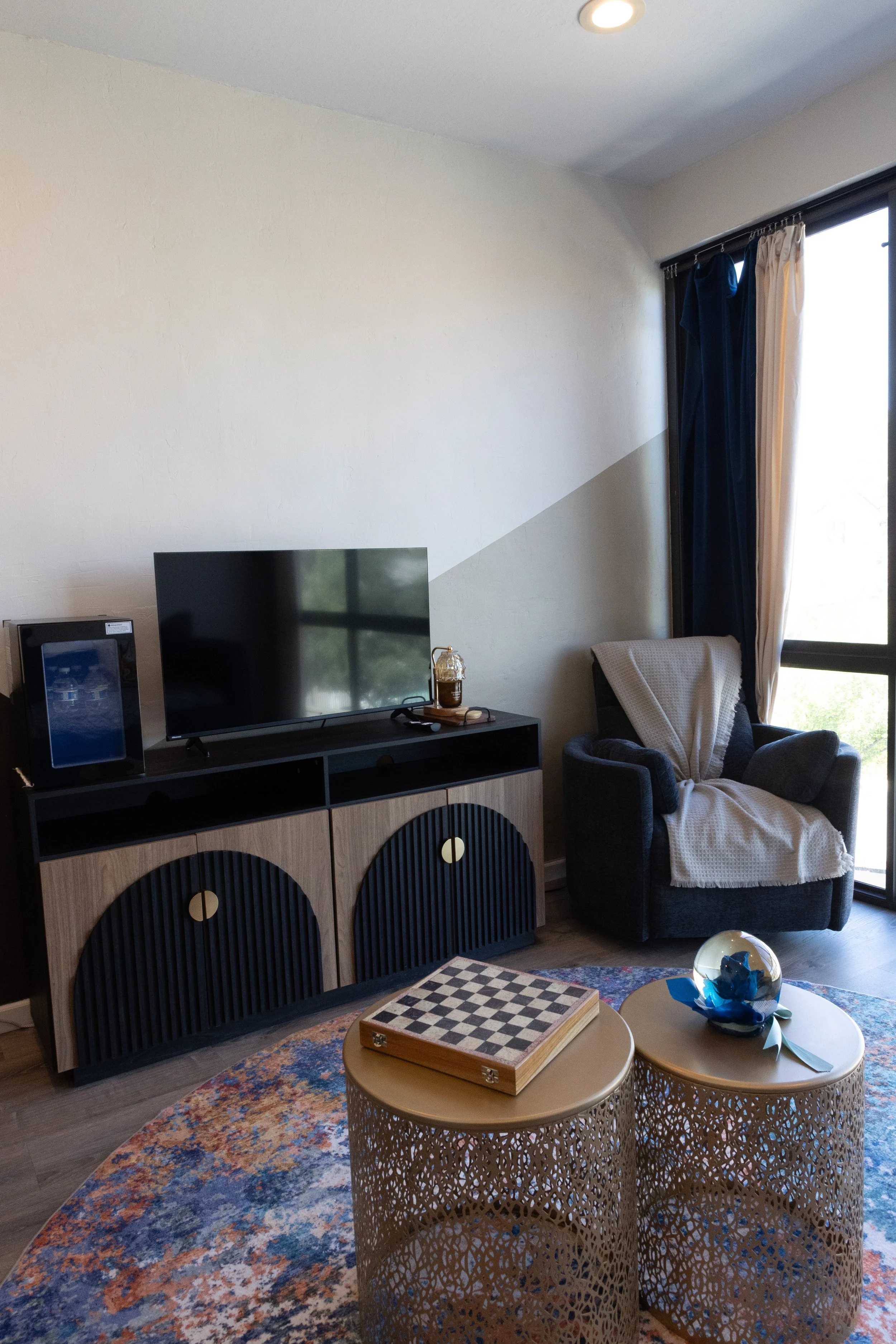 Living room with a large flat-screen TV, a small decorative owl statue on a stand, two round side tables with a chessboard and decorative blue glass art, an armchair with a white throw blanket, a colorful patterned rug, and a large window with curtai