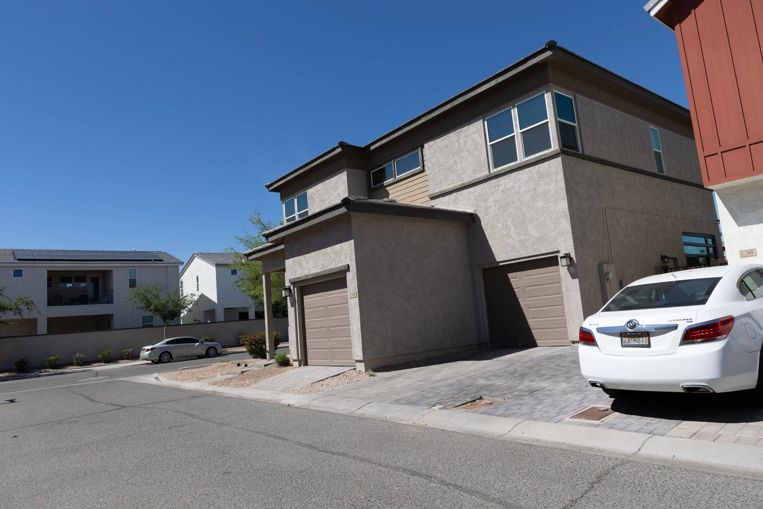 Residential street with modern houses, some with solar panels on the roofs, cars parked in driveways, and a clear blue sky.