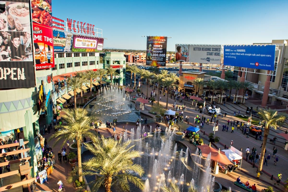 A busy outdoor shopping mall with palm trees, fountains, and large billboards advertising various stores and services, with many people walking around on a sunny day.
