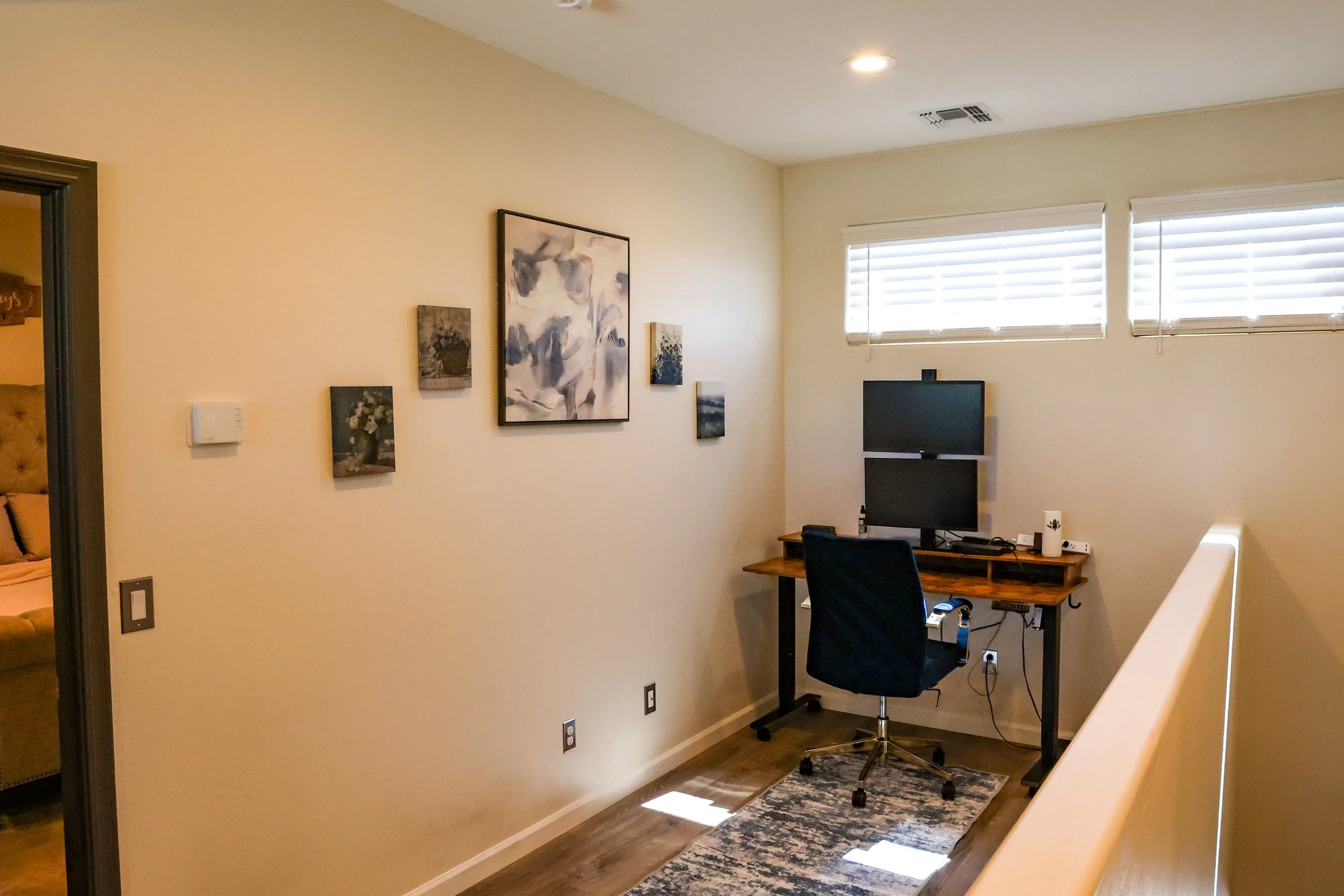 A home office corner with a wall decorated with five art pieces, a window with blinds, a computer monitor on a wooden desk, an office chair, a small rug, and a powered strip on the floor.