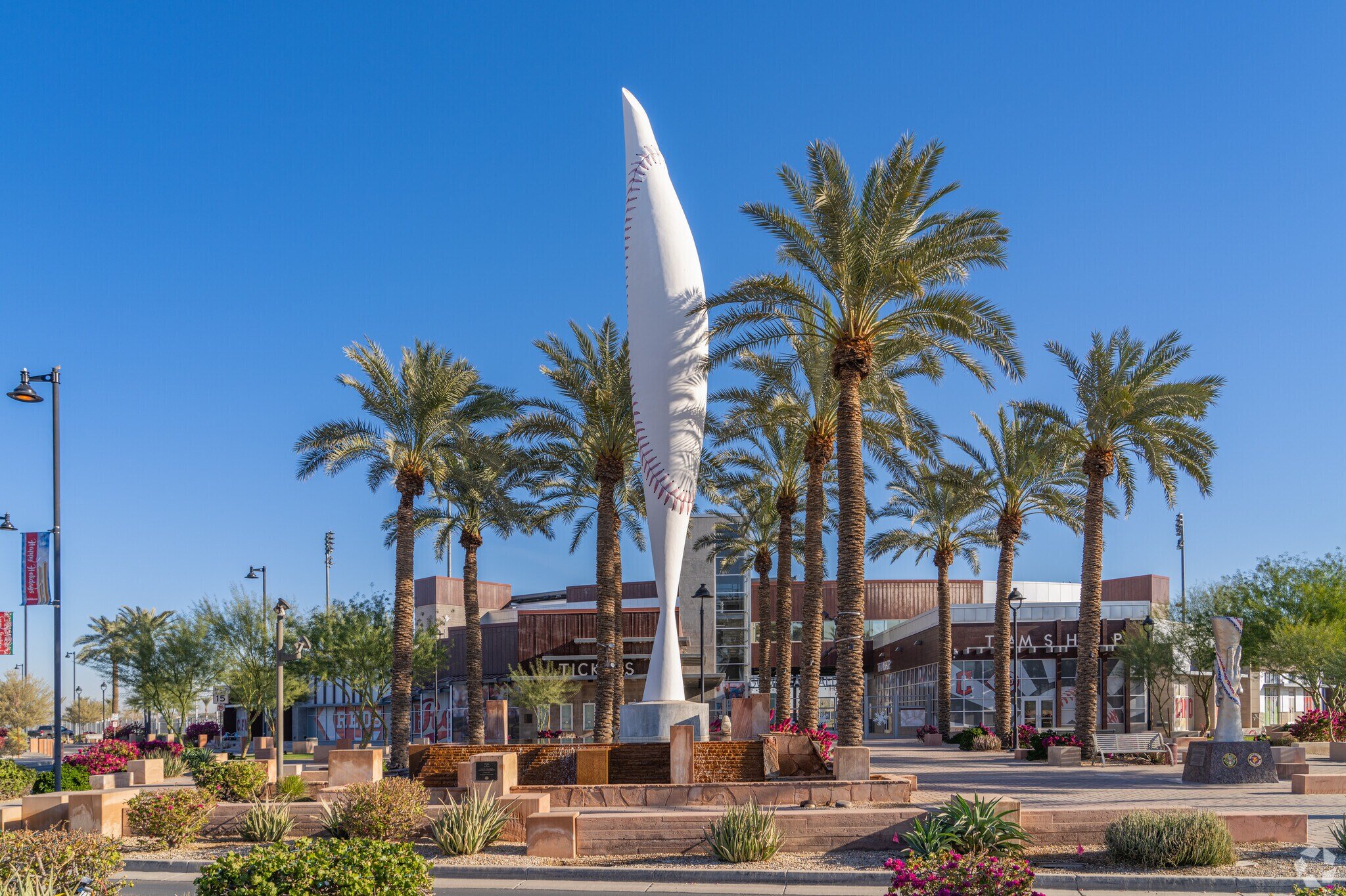Tall white sculpture that resembles a baseball bat or a wave, surrounded by tall palm trees in an outdoor plaza with benches, flowers, and a building in the background under a clear blue sky.