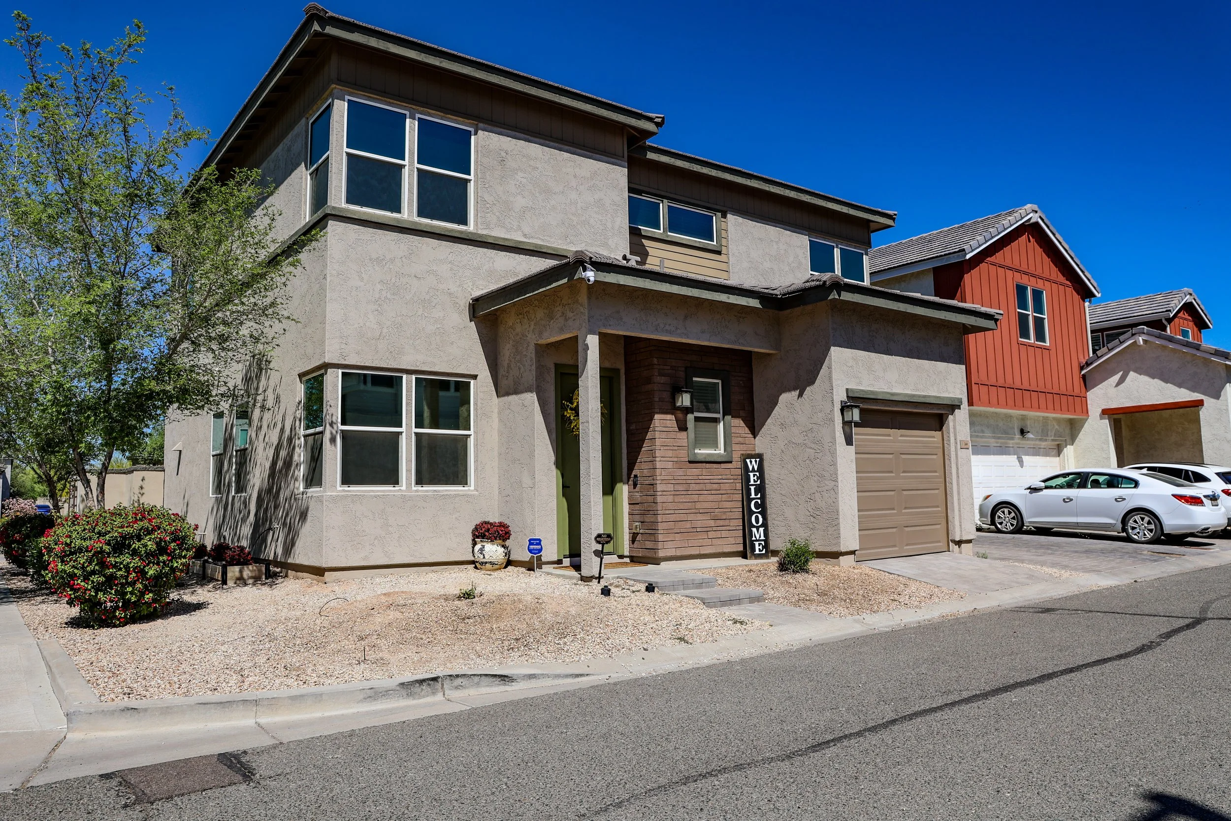 Front view of modern two-story house with beige and red exterior, featuring large windows, a garage, and a welcoming entrance with a vertical 'Welcome' sign, parked cars in driveway, surrounded by landscaping and a clear blue sky.