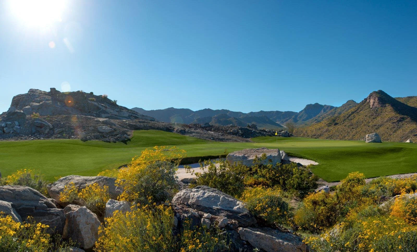 A scenic golf course is set against a backdrop of rocky mountains, with bright green grass, yellow flowering bushes, and a clear blue sky overhead.