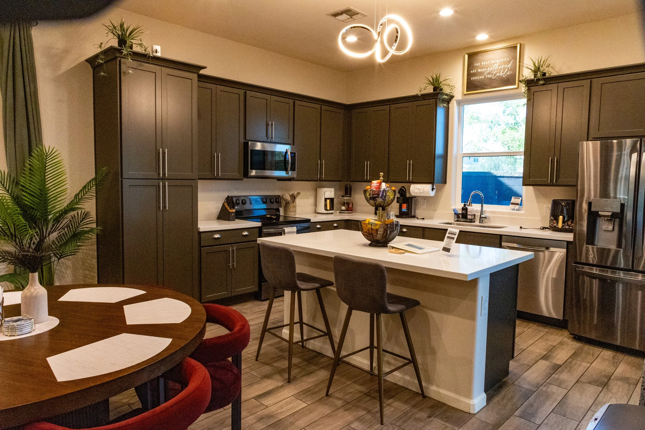 Modern kitchen with dark cabinets, stainless steel appliances, a white island with bar stools, and a dining table with red chairs. Decorative plants and a contemporary light fixture are also visible.