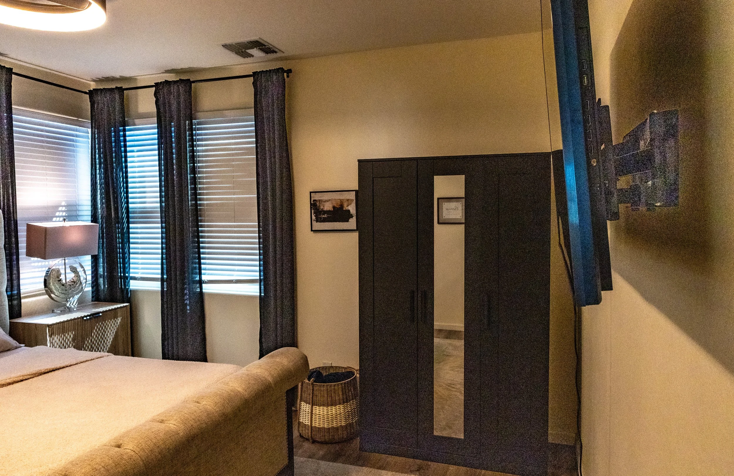 Living room with large windows covered by black curtains, a bed with beige bedding, a table with a lamp and decorative item, a dark dresser, and a TV against the wall.