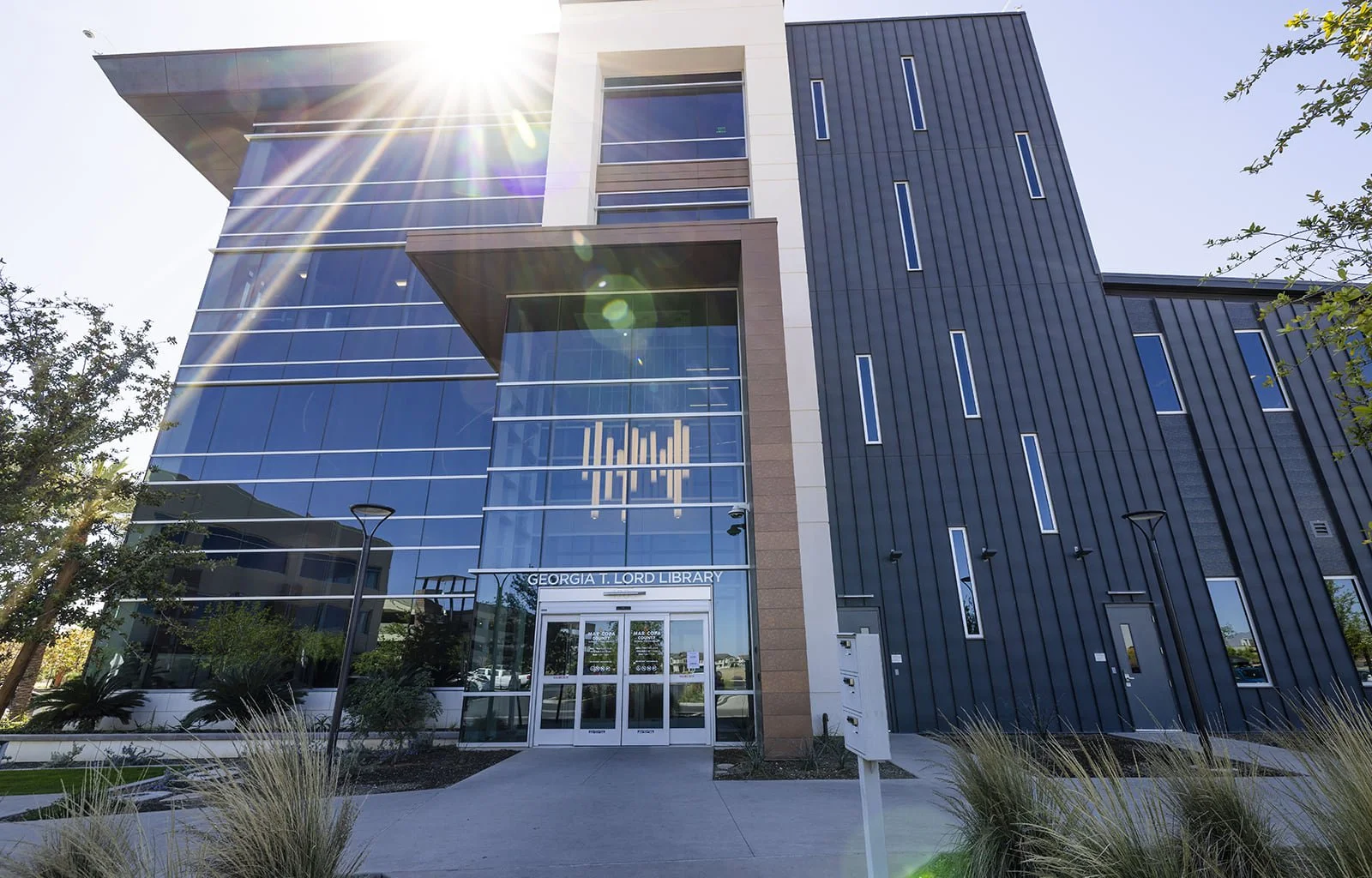 Modern multi-story library building with glass windows and blue accent panels, labeled as Georgia T. Lord Library, with landscaping and a clear sky.