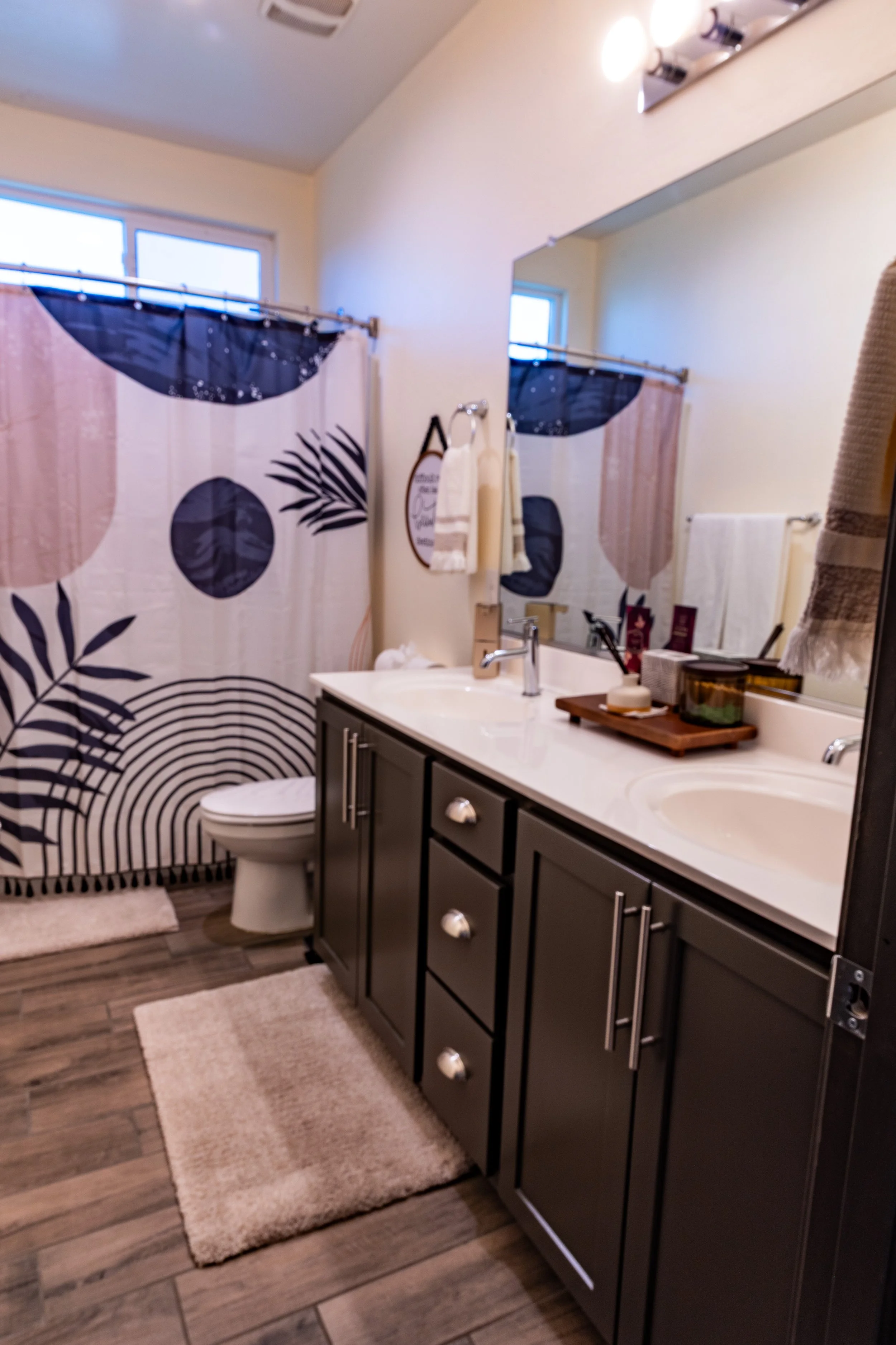 A bathroom with a dark gray vanity with silver handles, a white countertop with a sink, and a large mirror. There is a shower with a curtain featuring large black and beige leaves, a toilet, and a window letting in natural light. The floor is wood-lo