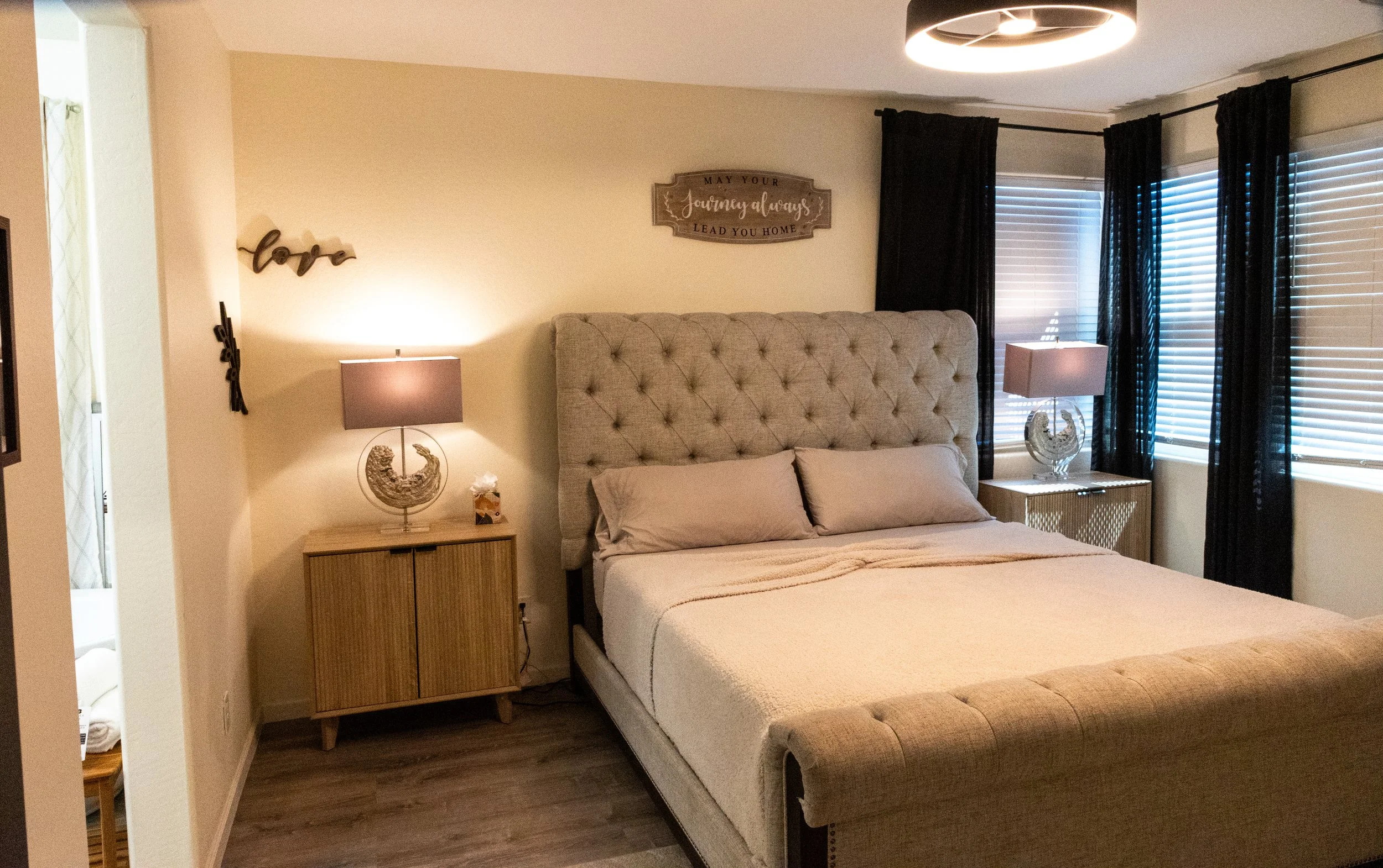 A bedroom with a large beige upholstered bed, two bedside tables with lamps, dark curtains over windows, and wall decorations including a wooden sign and a wall-hanging with the word 'love'.
