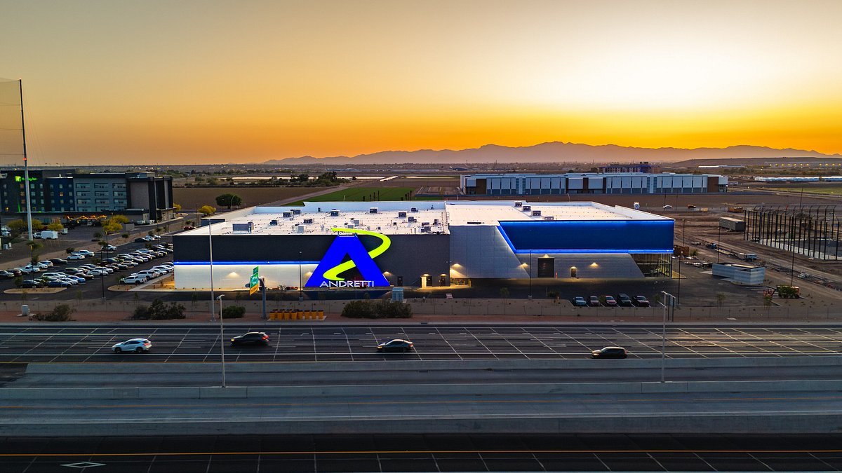 A large commercial building with a blue, white, and black exterior, illuminated by bright lights, at sunset. The building features a prominent logo with a blue and green swoosh and the word "Andretti." In the foreground, there is a highway with a few