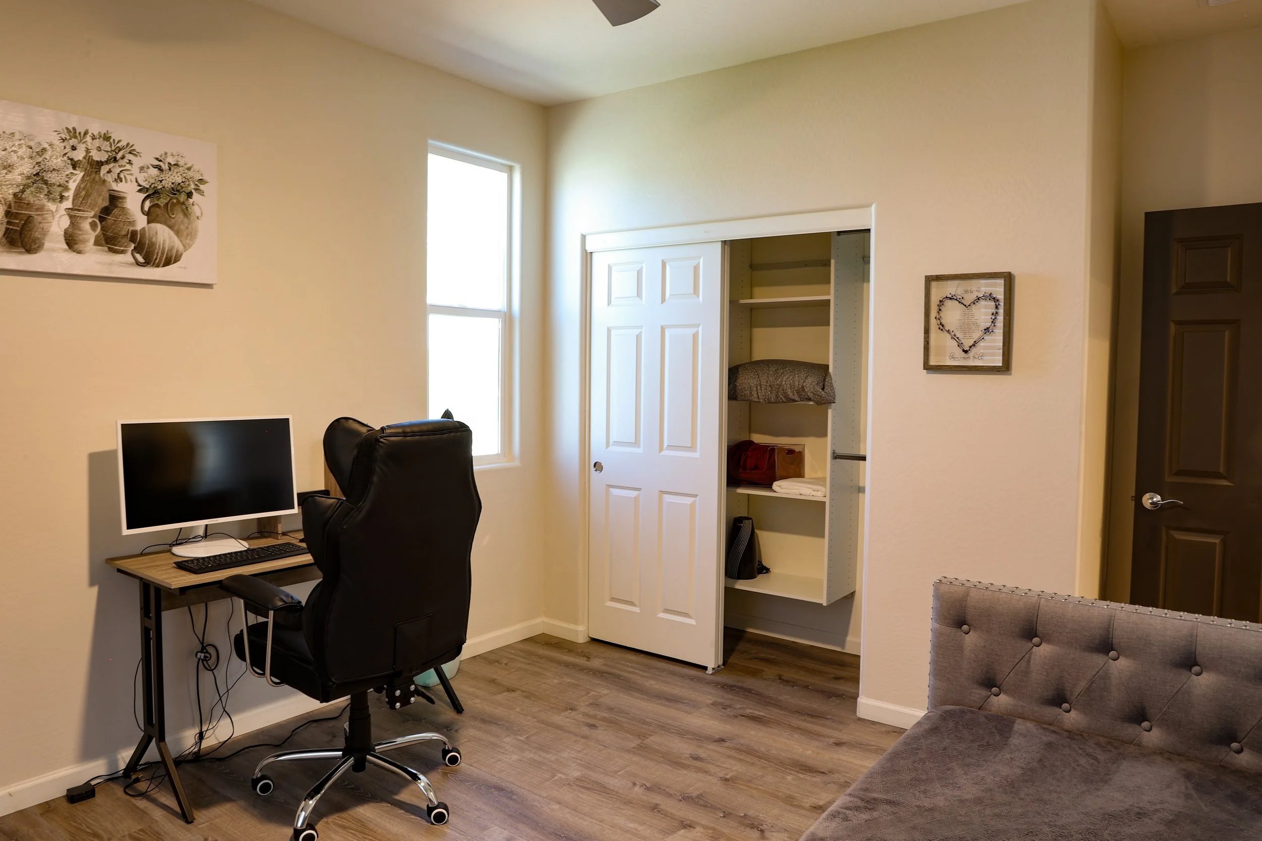 A cozy home office with a desk, a computer, a black office chair, a small artwork with a heart made of flowers, a closet with shelves holding folded clothes, a window, and a brown door. The room has beige walls and wood flooring.