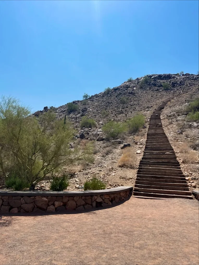 Wooden staircase leading up a rocky desert hillside with sparse vegetation, under a clear blue sky.