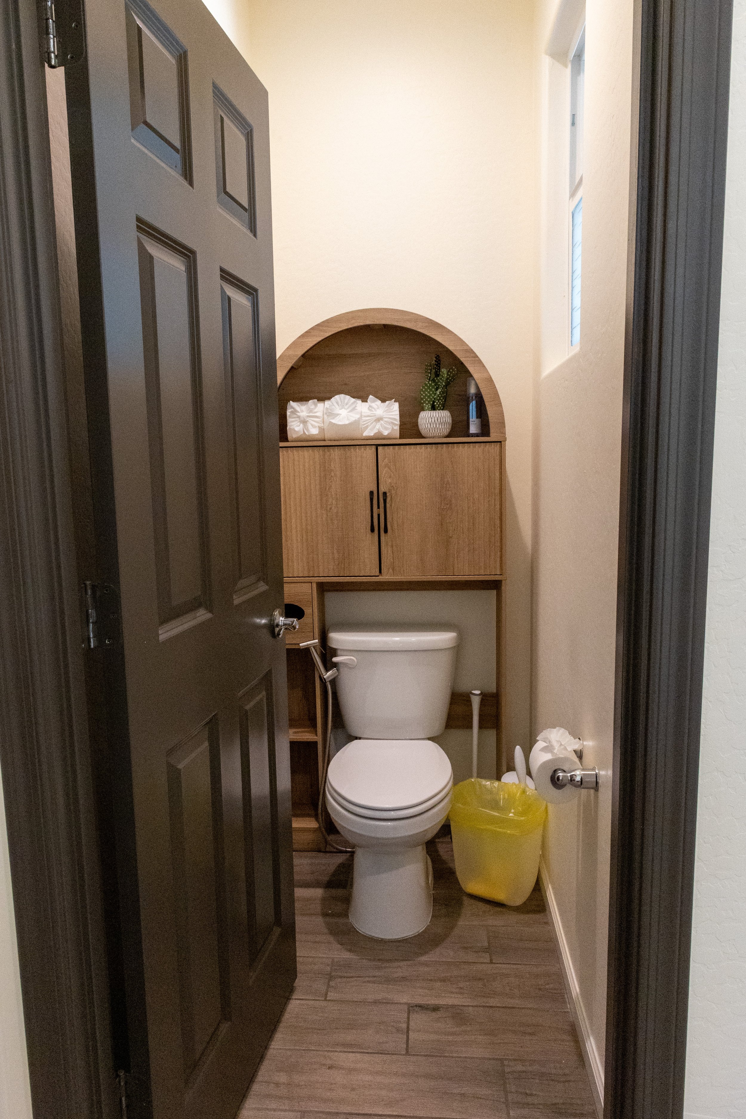 A small bathroom with a gray door, a toilet, a wooden cabinet with drawers and a curved top, a small potted cactus, a tissue box, a blue bottle, a yellow trash bin, and a window with white blinds.