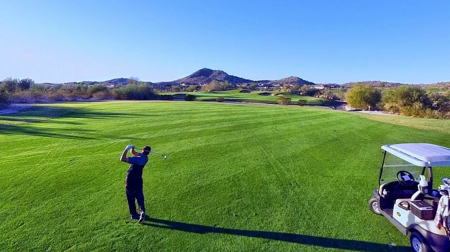 Golfer swings a club on a lush golf course with a golf cart nearby, landscape with hills and trees in the background.